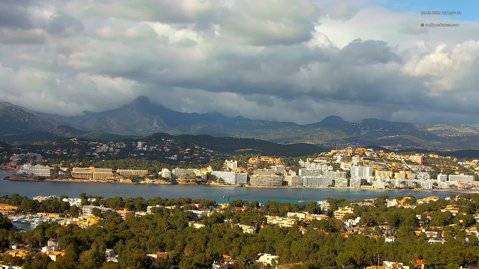 A coastal town nestled between a calm bay and a mountain range under a partly cloudy sky.