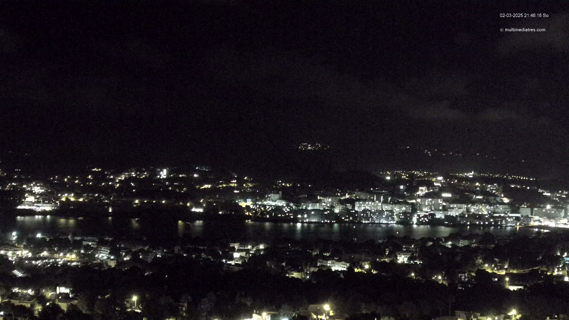 A nighttime view of a city with a body of water, under a dark, mostly cloudy sky.