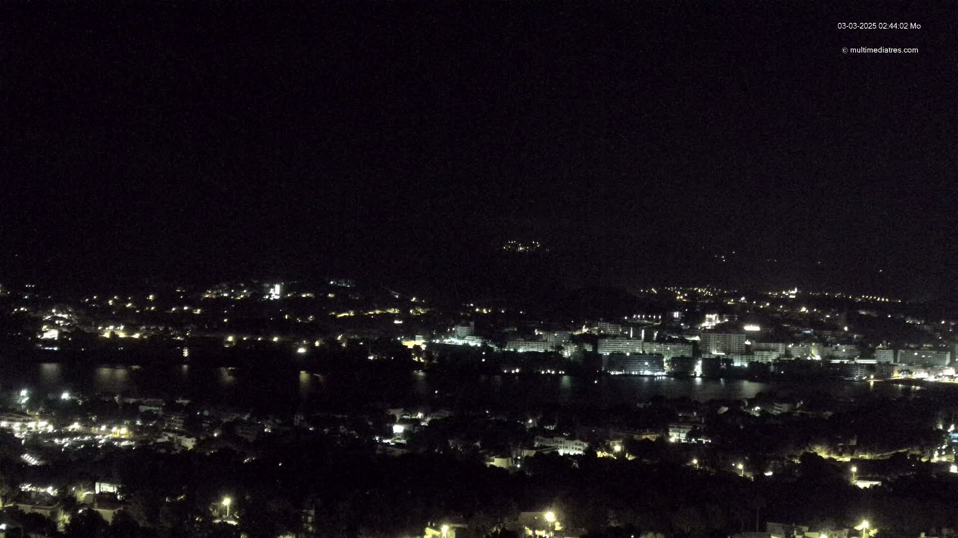 A nighttime view of a city and body of water illuminated by scattered lights under a clear, dark sky.