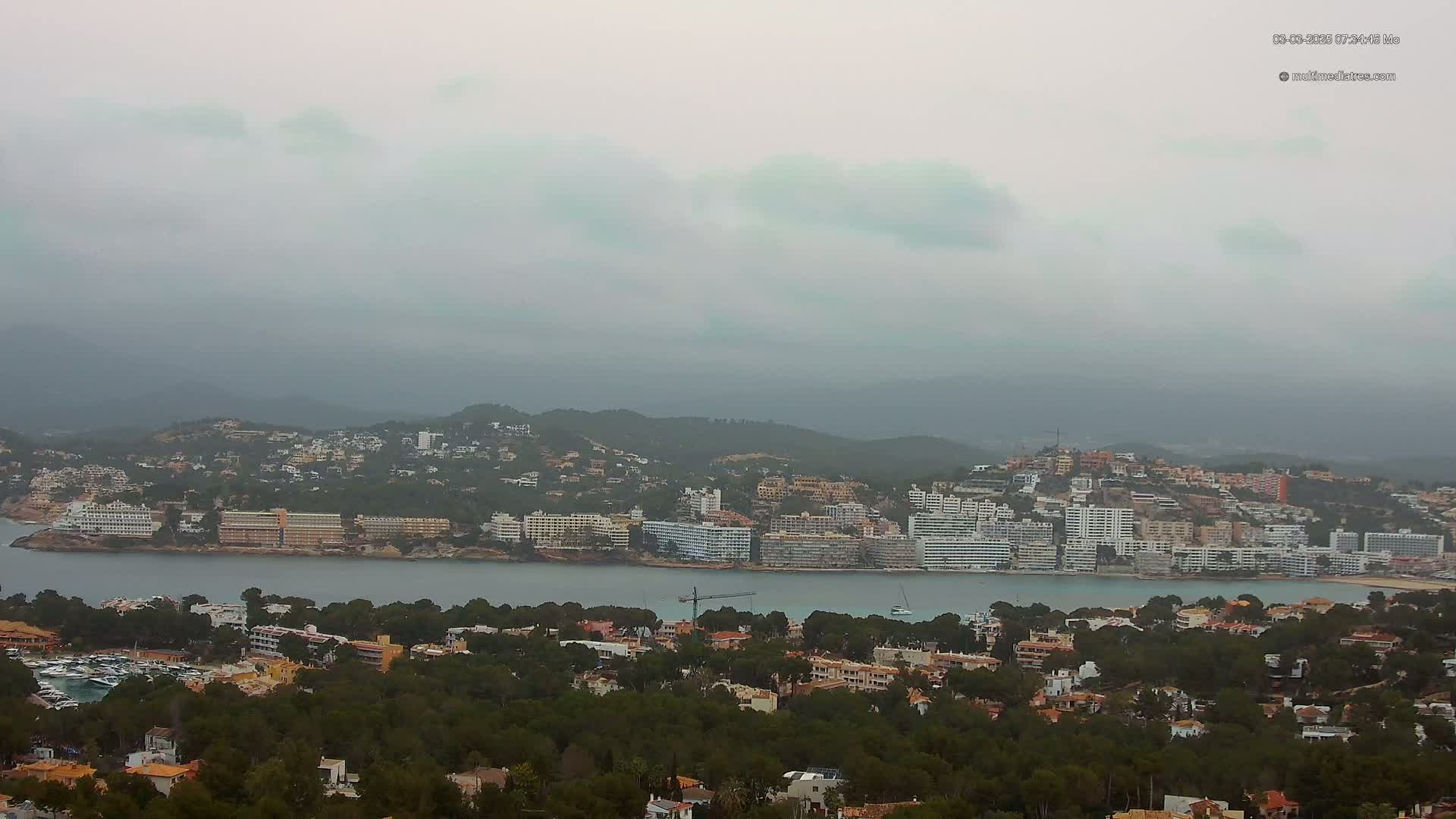 A coastal town nestled in a bay, surrounded by hills and lush greenery, under an overcast sky.