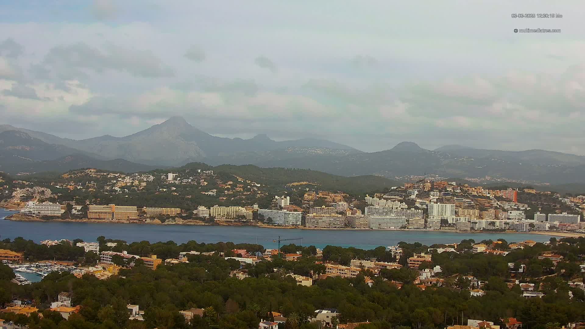 A coastal town nestled amongst green hills and mountains under an overcast sky overlooks a calm bay.
