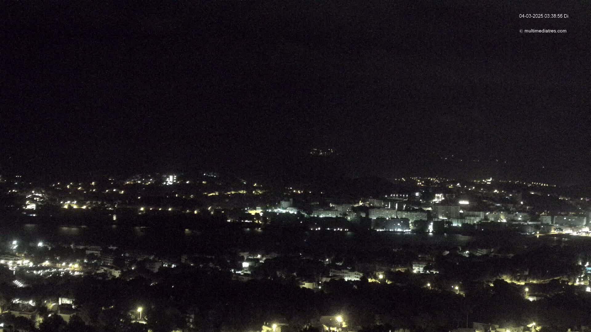 A nighttime, long-range view of a coastal town with scattered lights, situated near a body of dark water, under a clear, dark sky.