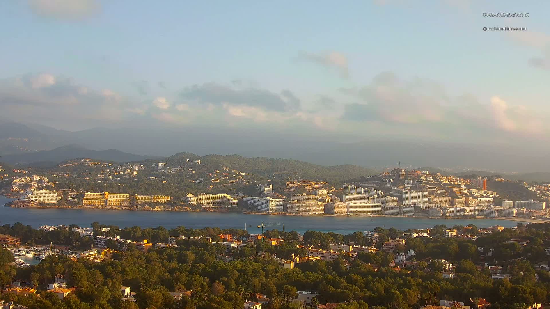 A coastal town nestled in a hilly landscape under a partly cloudy sky, with calm waters separating the town from a shoreline lined with hotels.