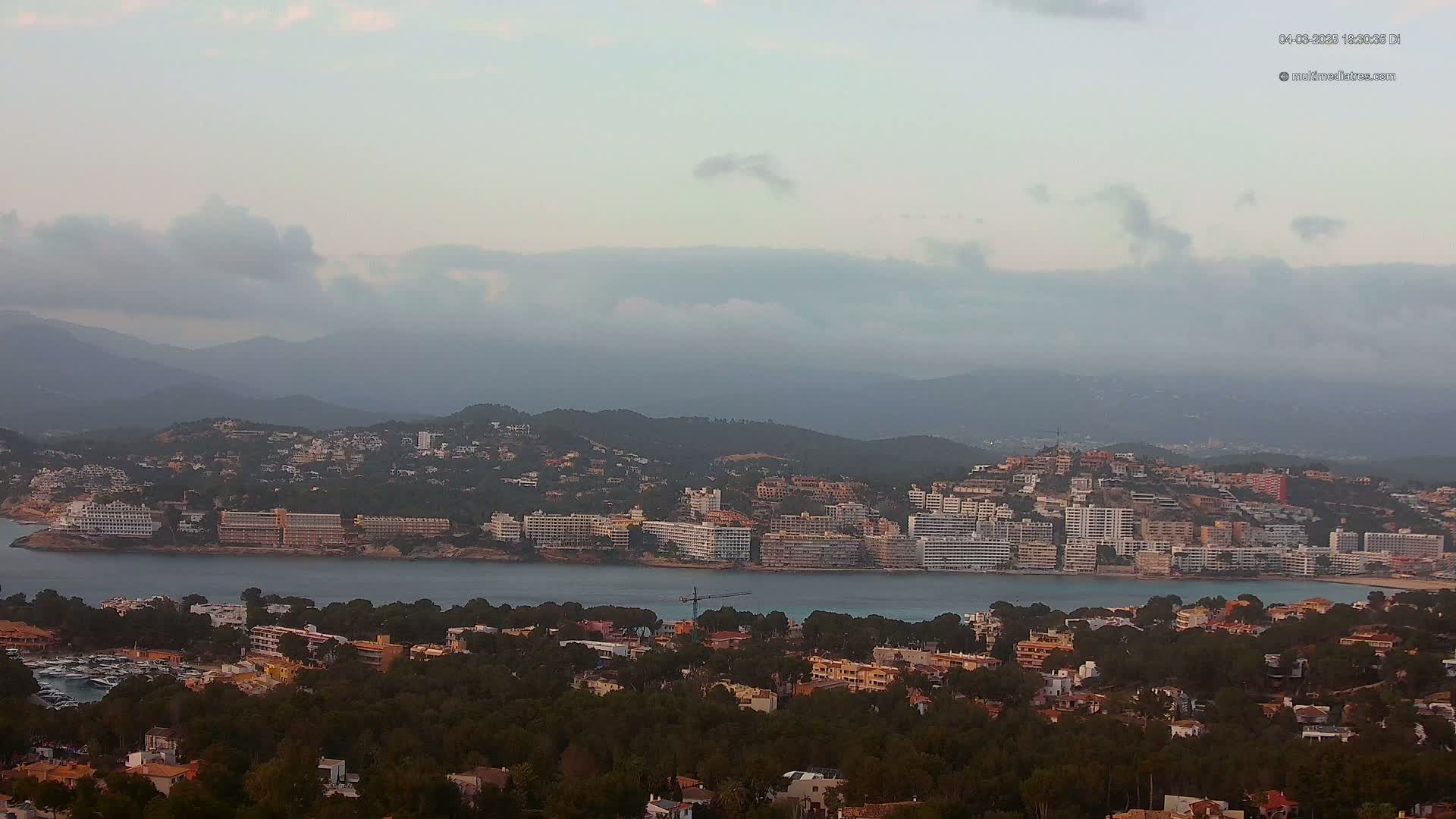 A coastal town with numerous buildings sits nestled between a body of water and a mountainous backdrop under a cloudy sky.