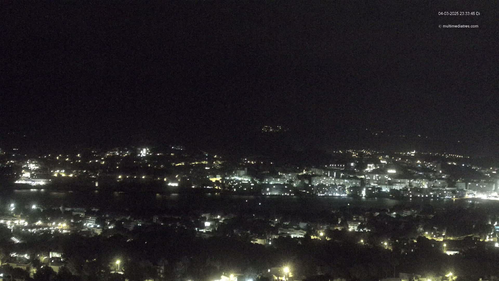 A nighttime, long-range view of a coastal city illuminated by scattered lights, situated beside a dark body of water under a clear, dark sky.