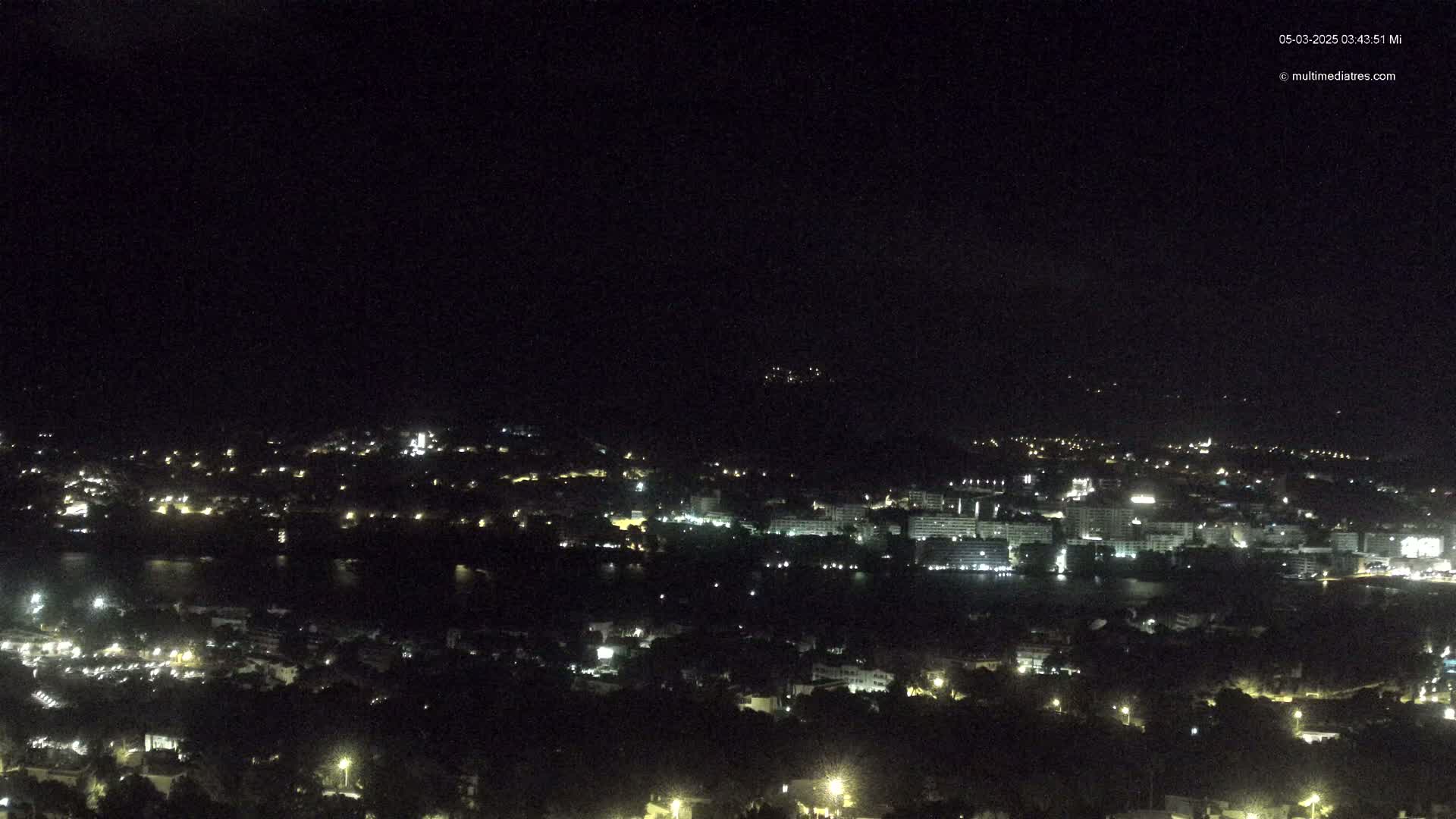 A nighttime, high-angle view shows a coastal town with scattered lights and a body of dark water.