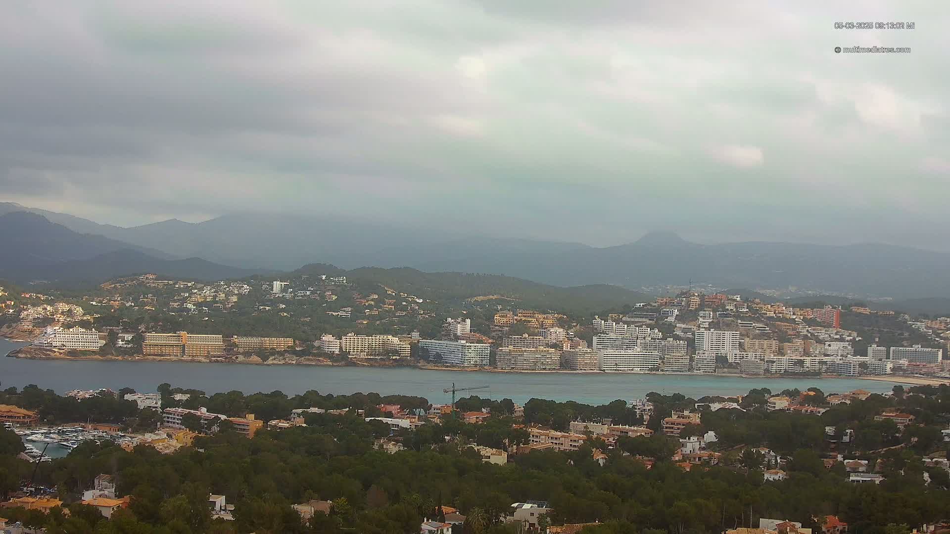 A coastal town nestled among green hills and mountains under an overcast sky, with a calm body of water separating the town from a line of hotels.