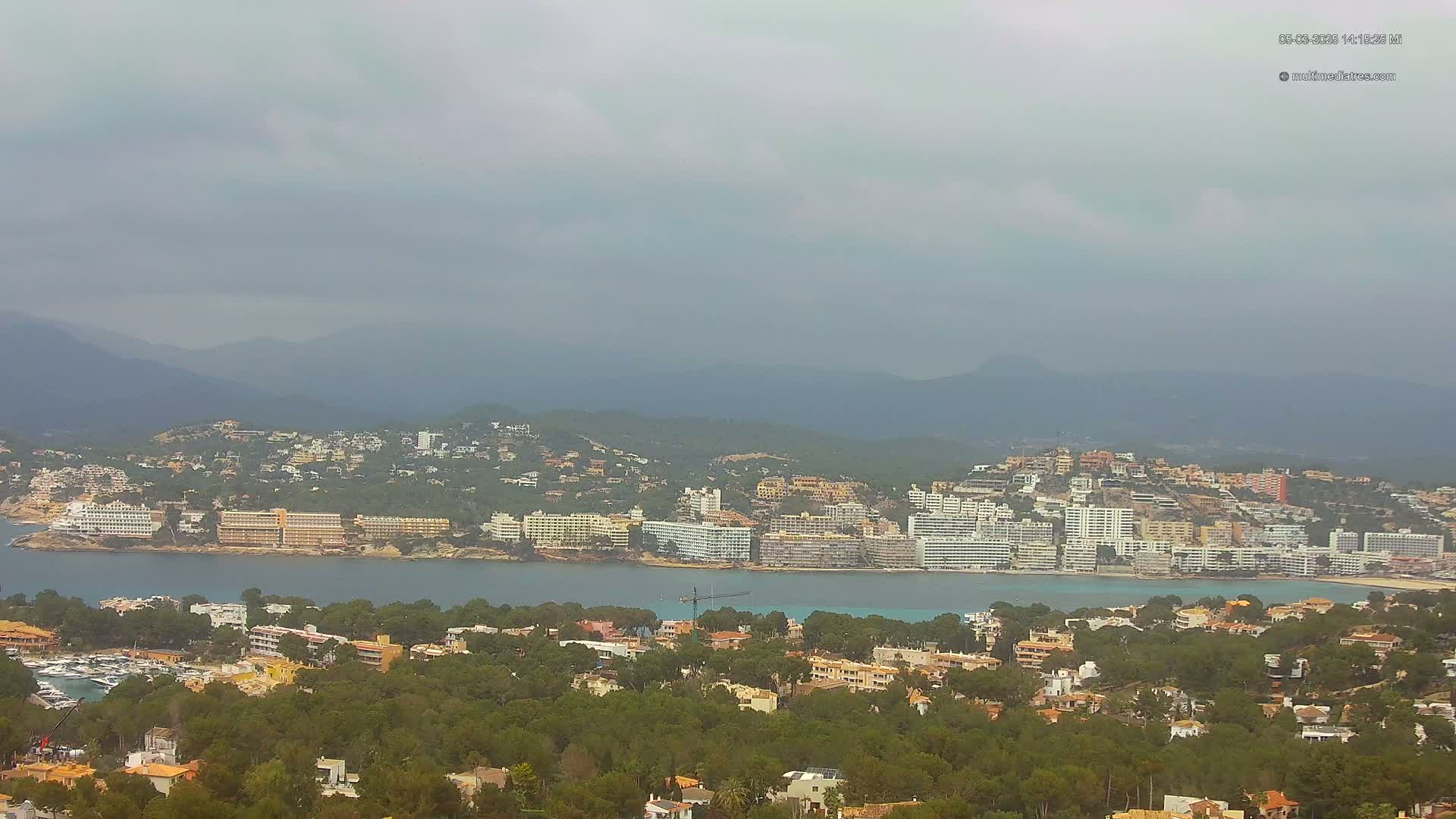 A coastal town nestled in a bay, surrounded by green hills and mountains under an overcast sky.