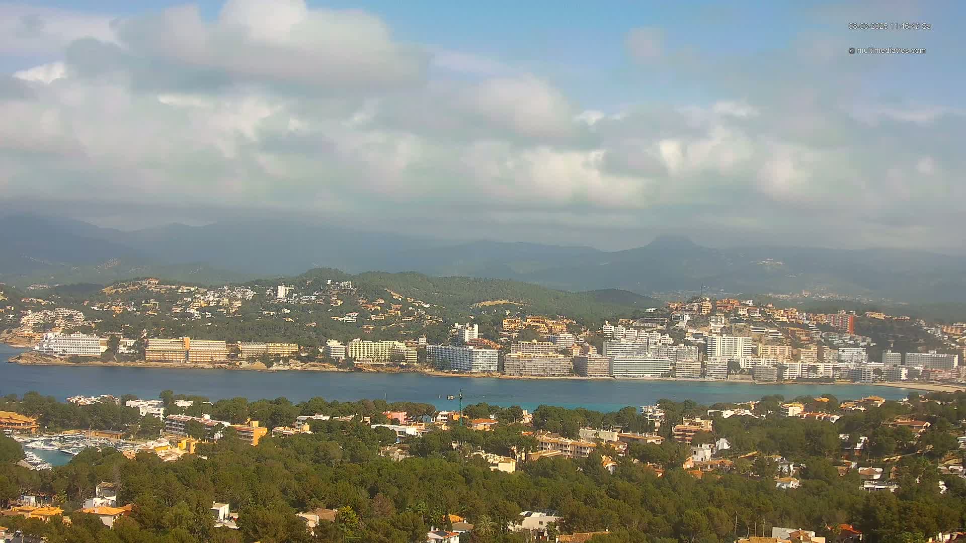 A coastal town nestled among green hills and a calm bay under a partly cloudy sky.