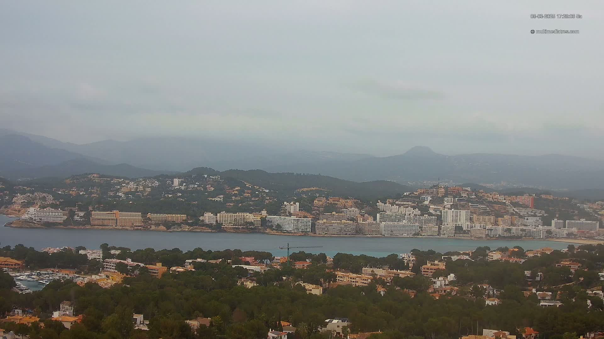 A coastal town nestled between a body of water and hazy mountains under an overcast sky.