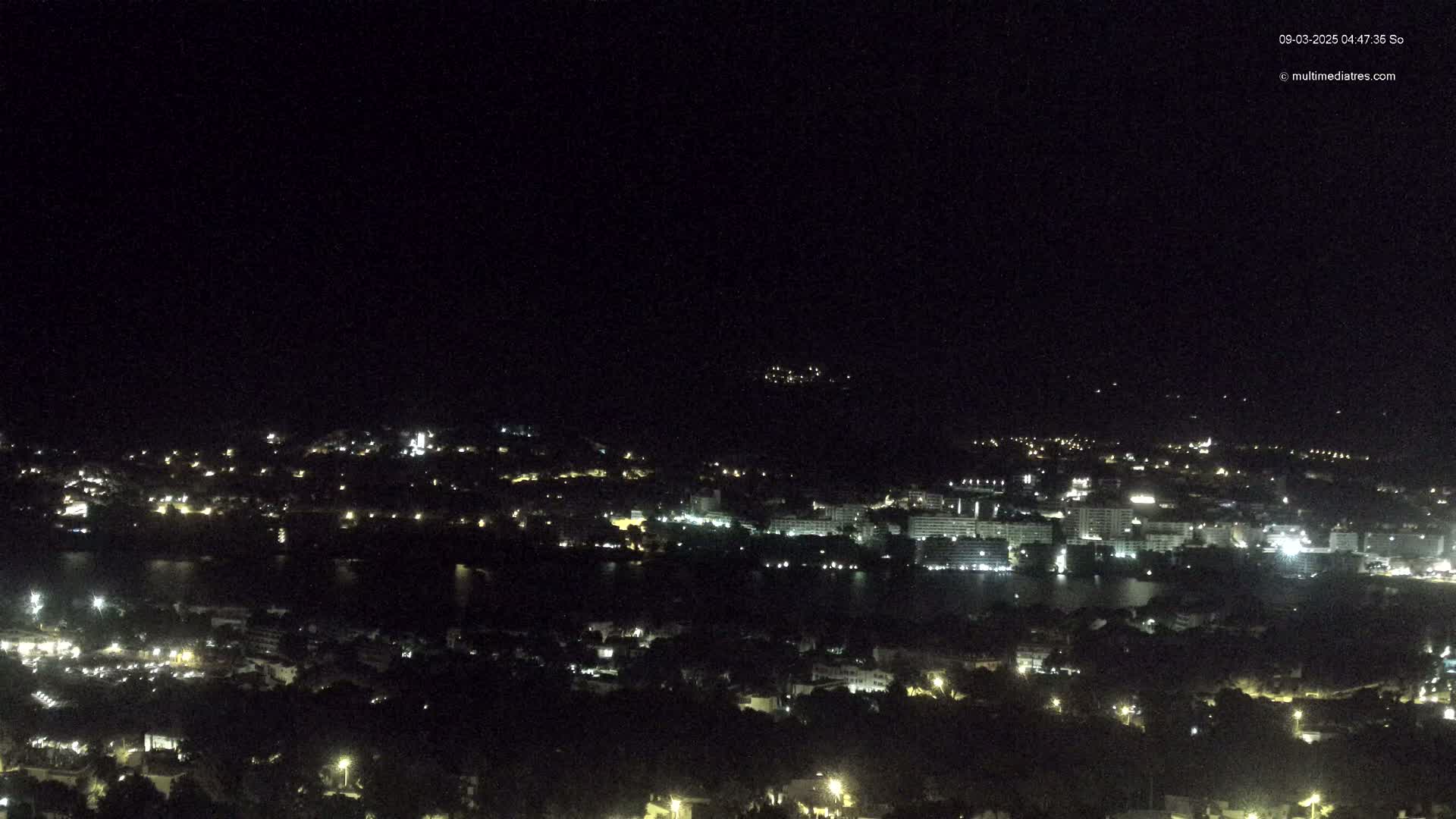 A nighttime, high-angle view shows a coastal town with many lights near a dark body of water, under a clear, dark sky.