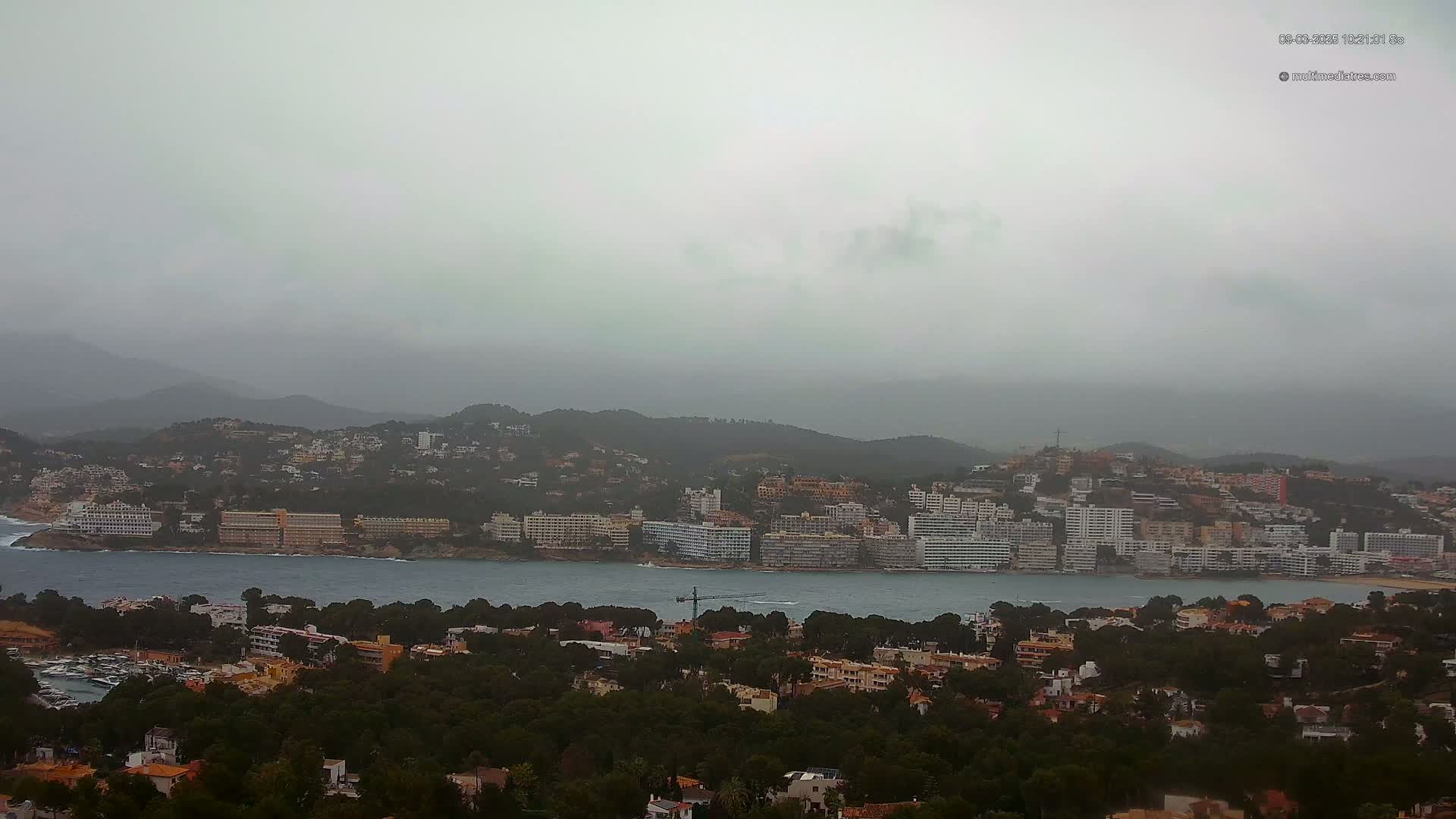 A coastal town nestled amongst hills and a body of water under an overcast sky.