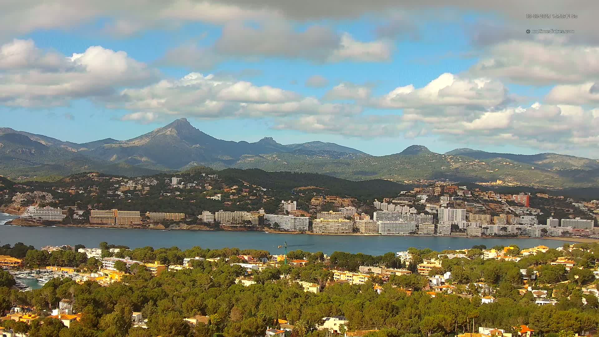 A coastal town with numerous buildings sits nestled amongst green hills and a calm bay, with mountains in the background under a partly cloudy sky.
