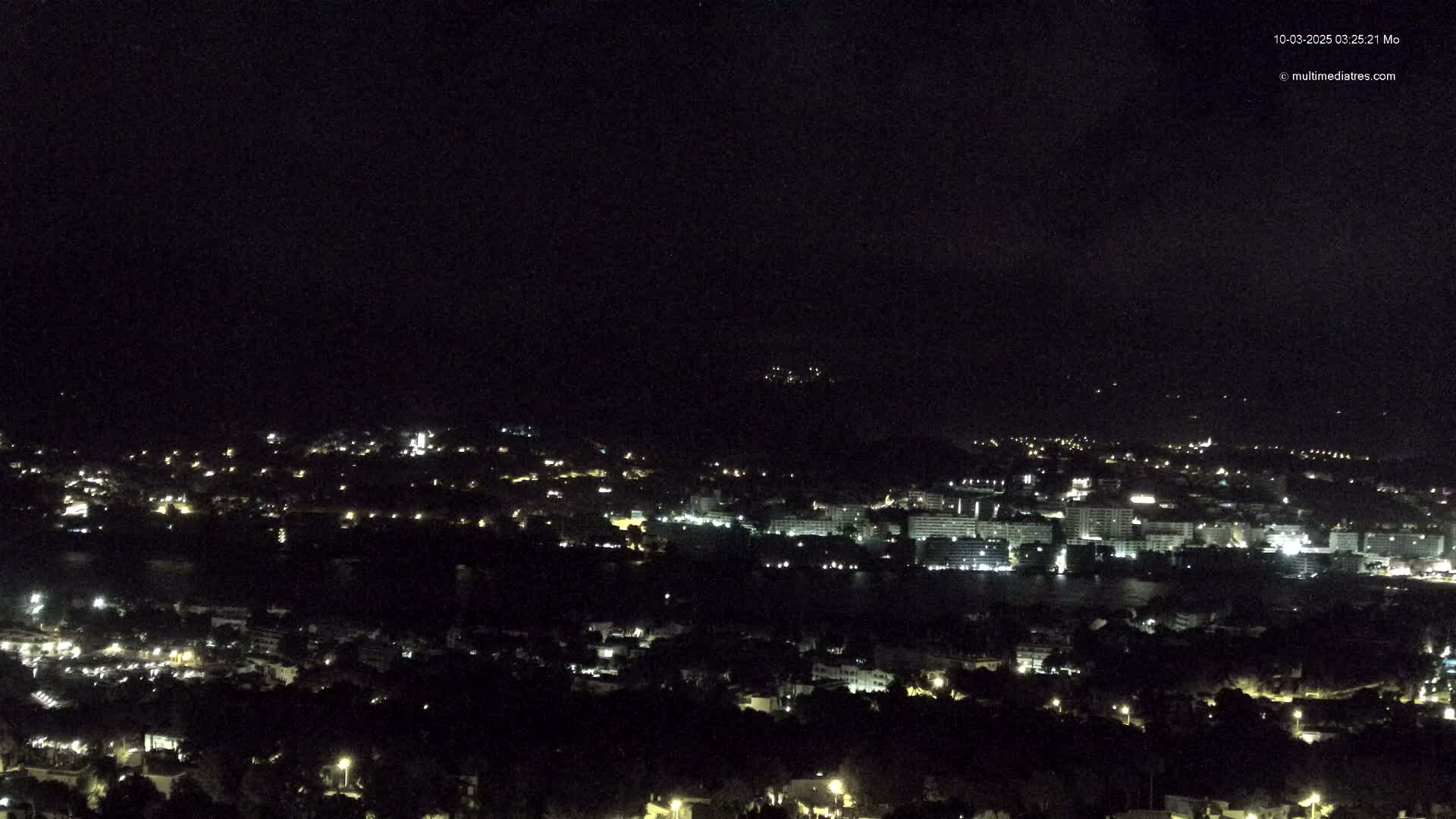 A nighttime, high-angle view of a coastal city with numerous lights illuminating buildings and streets, under a dark, clear sky.