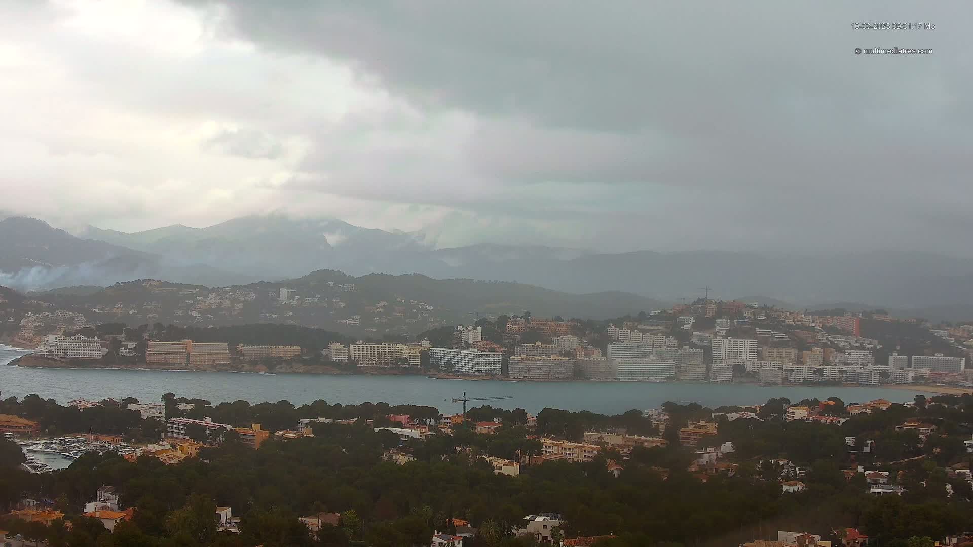 A coastal town nestled among dark green hills and a calm bay under an overcast sky.