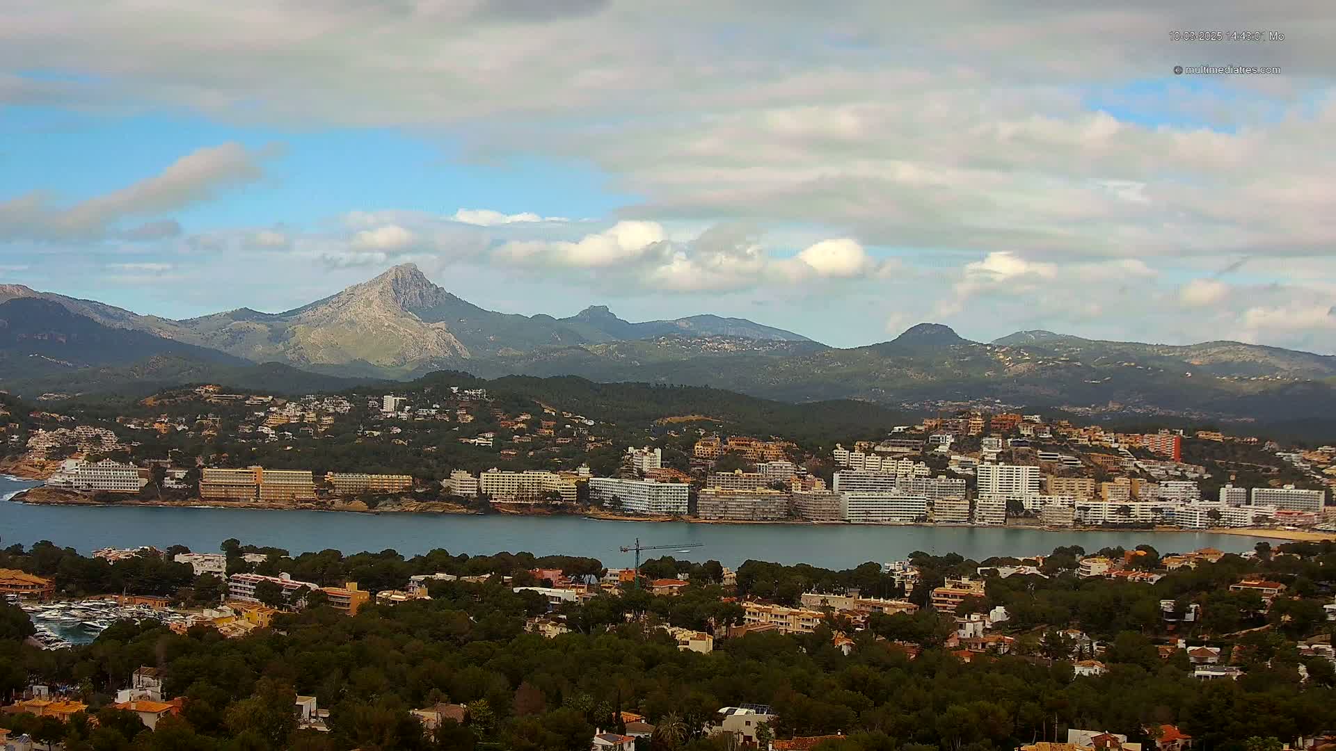 A coastal town nestled between a calm bay and a mountain range under a partly cloudy sky.