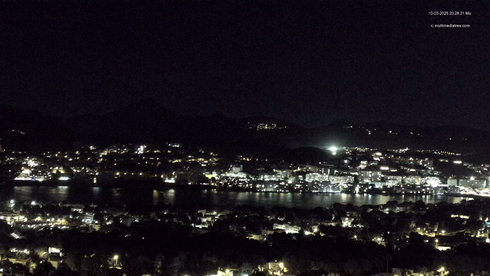 A nighttime view of a coastal city with its lights illuminating the buildings and water, under a clear, dark sky.