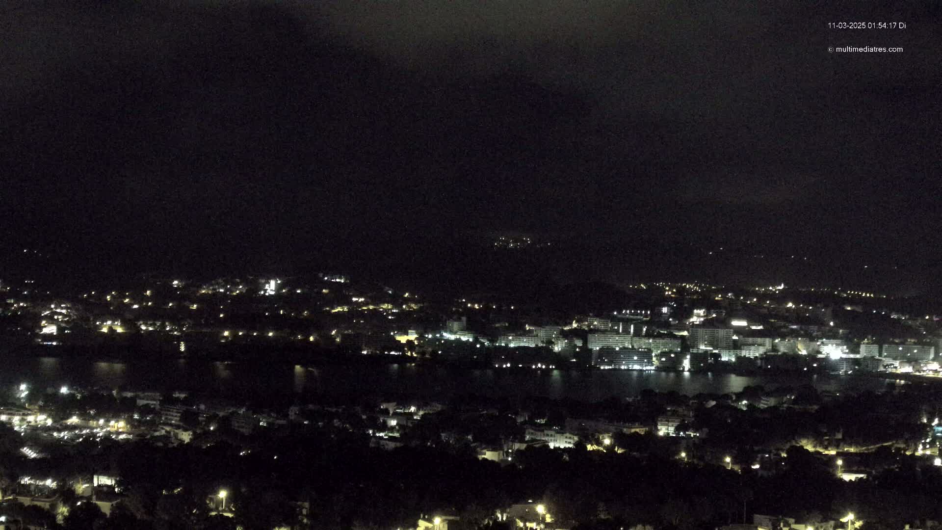A nighttime view of a coastal city with a body of water, under a dark, cloudy sky.