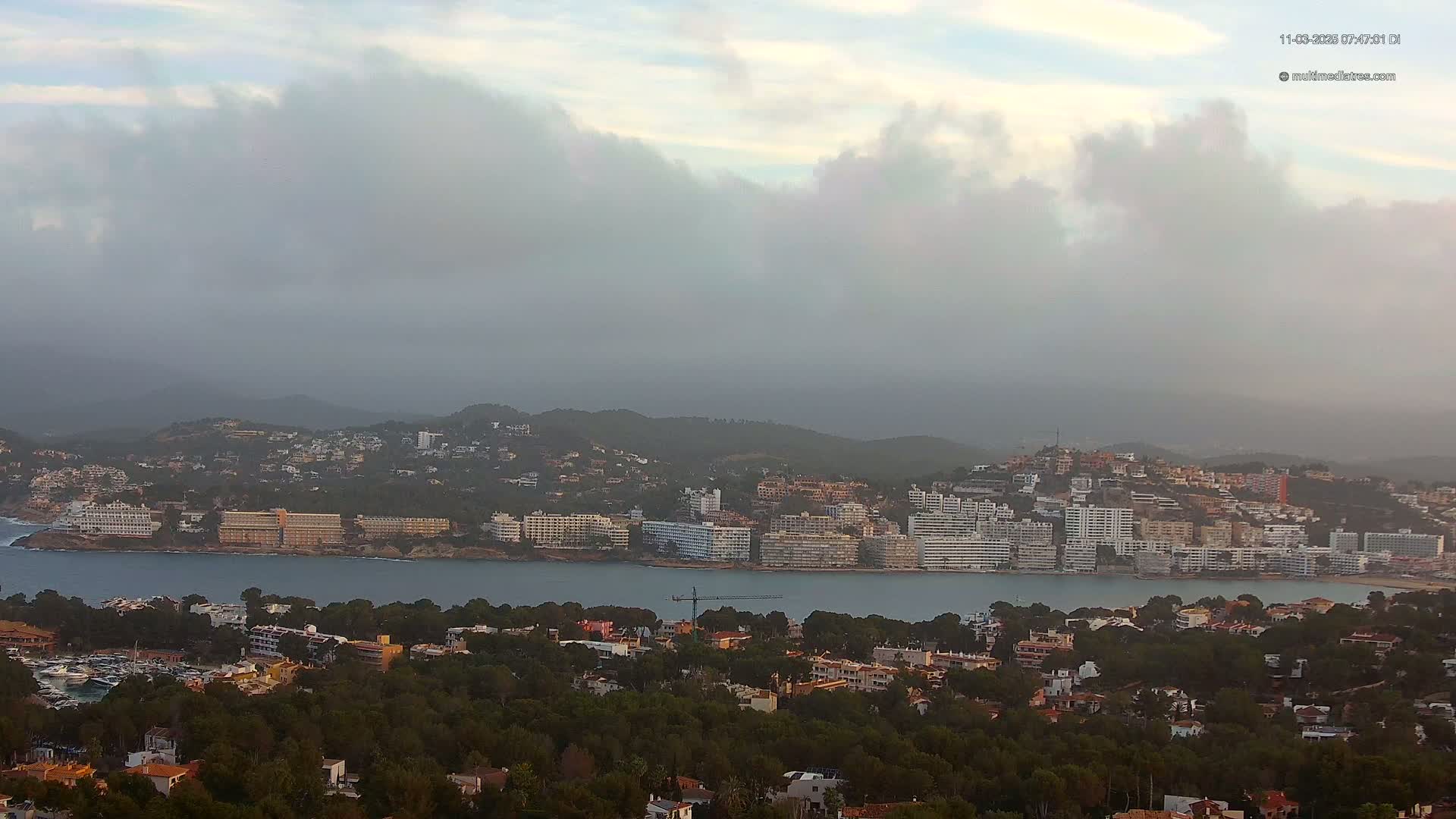 A hazy coastal town nestled among hills, with a body of water separating a line of hotels from a densely wooded lower area.