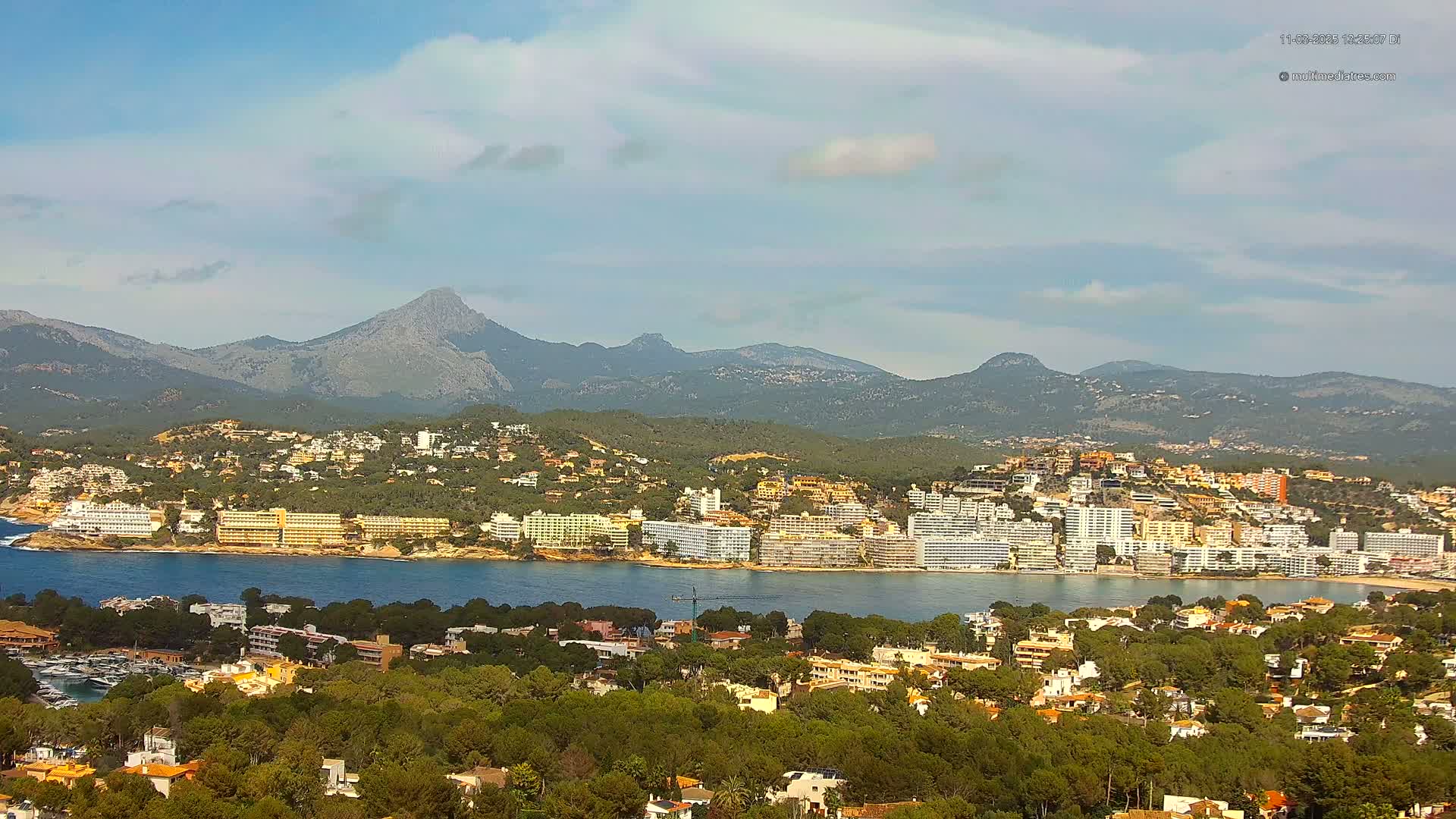 A coastal town with numerous buildings is nestled at the base of a mountain range under a partly cloudy sky.