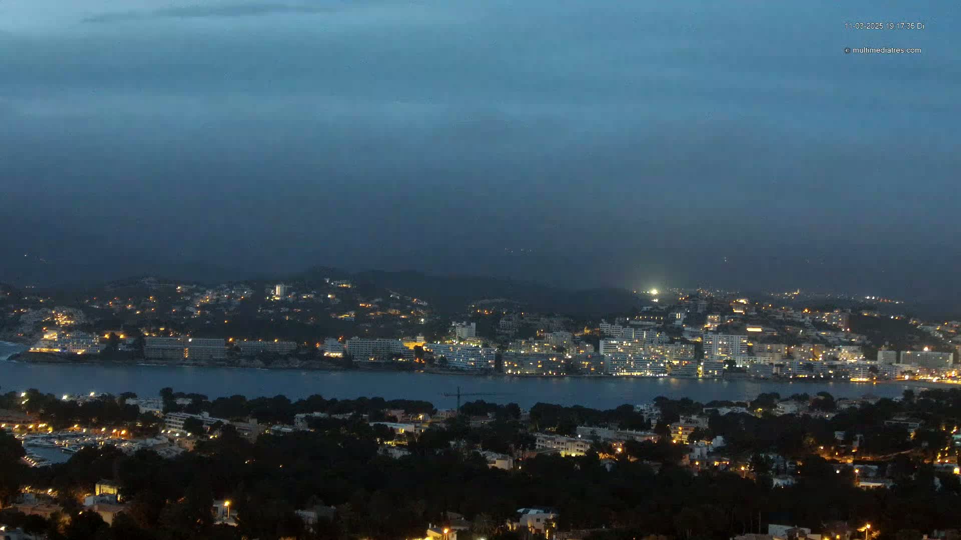 A coastal town at night, with its lights twinkling against a dark, overcast sky.
