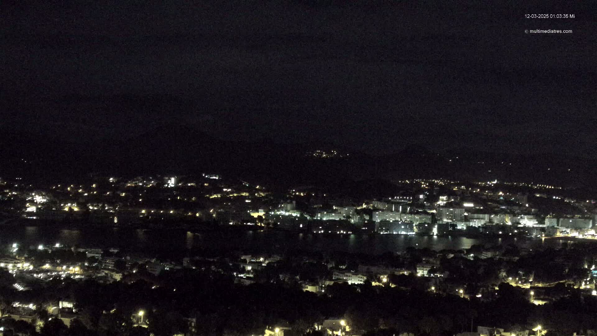 A nighttime, aerial view shows a coastal city with many lights scattered across the landscape,  illuminating buildings near a dark body of water under a clear, dark sky.