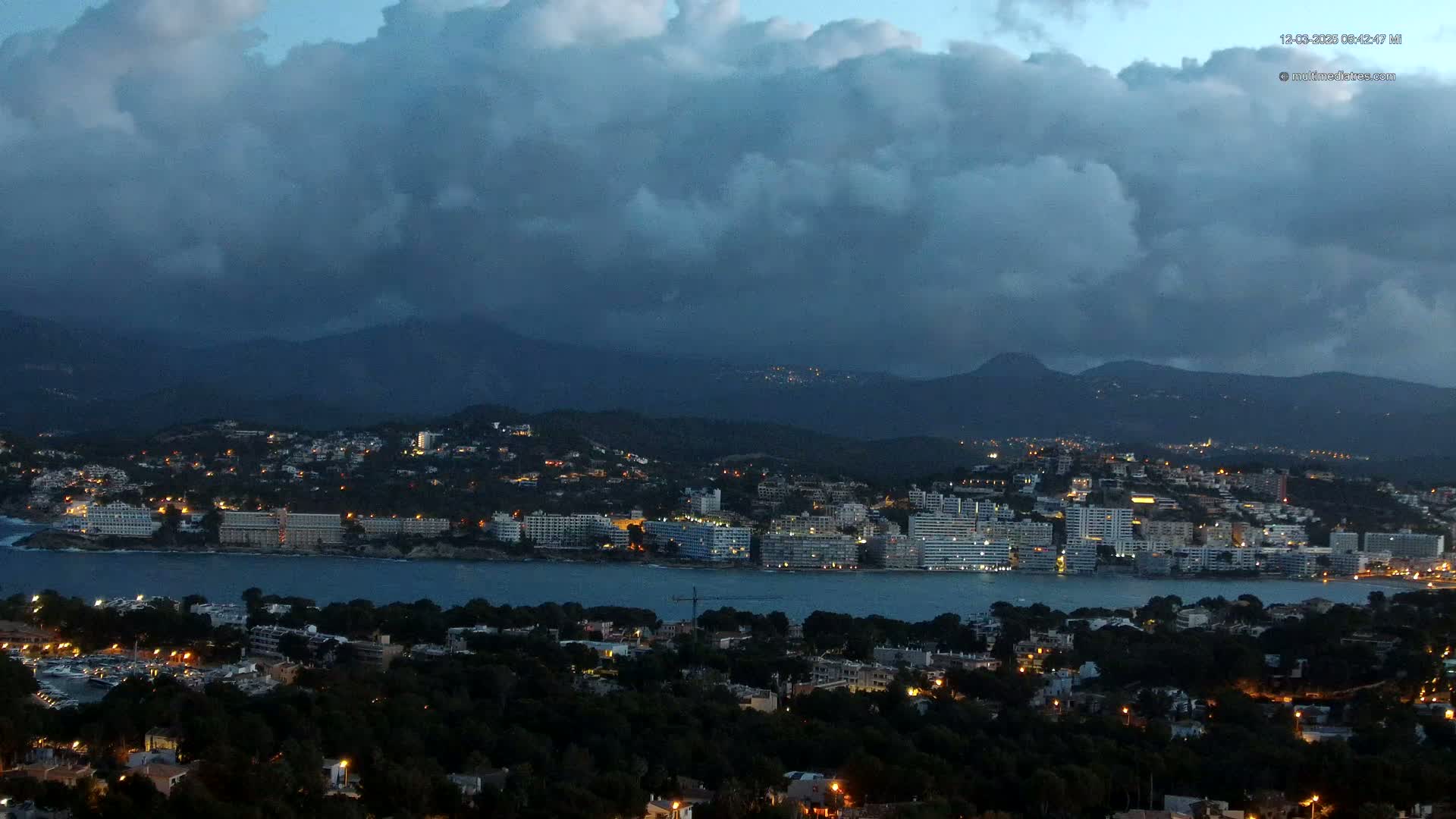A coastal town with many lights is nestled between a body of water and dark, cloud-covered mountains at twilight.