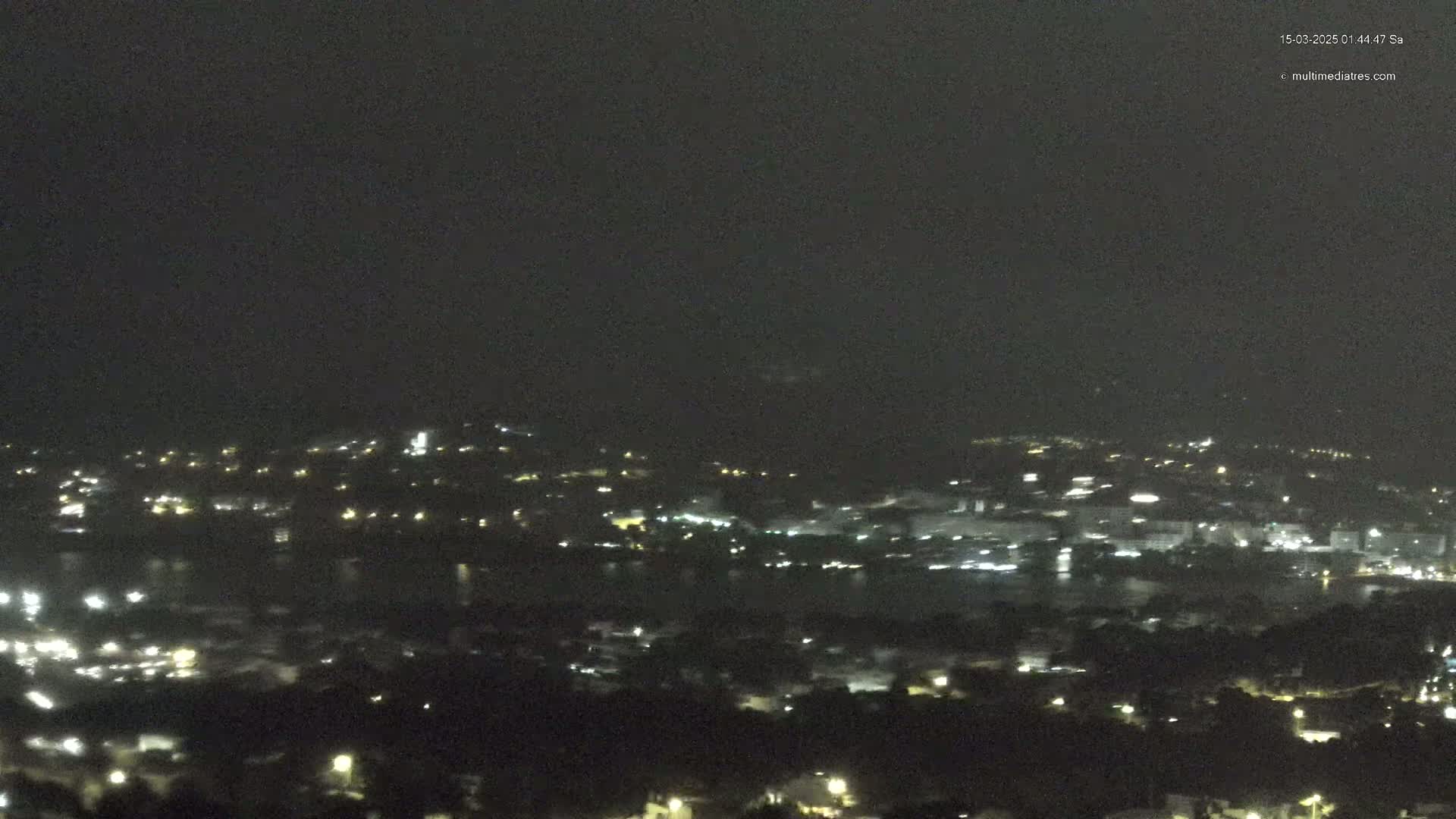 A nighttime, high-angle view shows a city illuminated by scattered lights alongside a dark body of water.