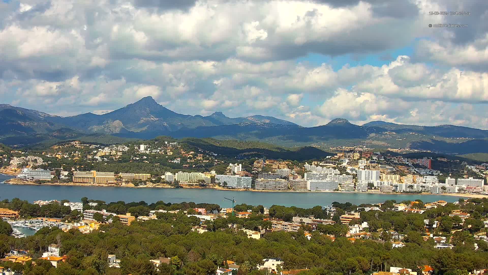 A coastal town nestled beside a calm bay, with hotels and residential buildings overlooking the water, is set against a backdrop of mountains under a partly cloudy sky.