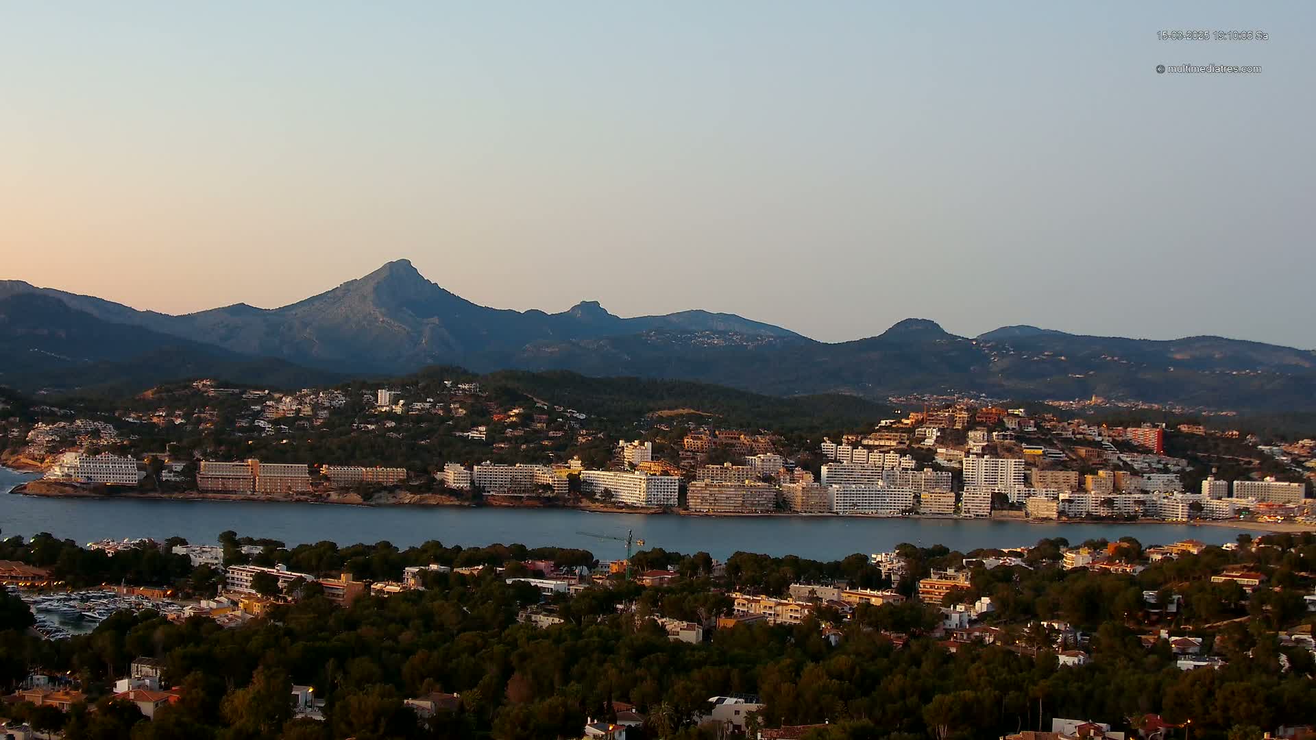 A coastal town nestled between a calm bay and a mountain range under a clear, pale sky.