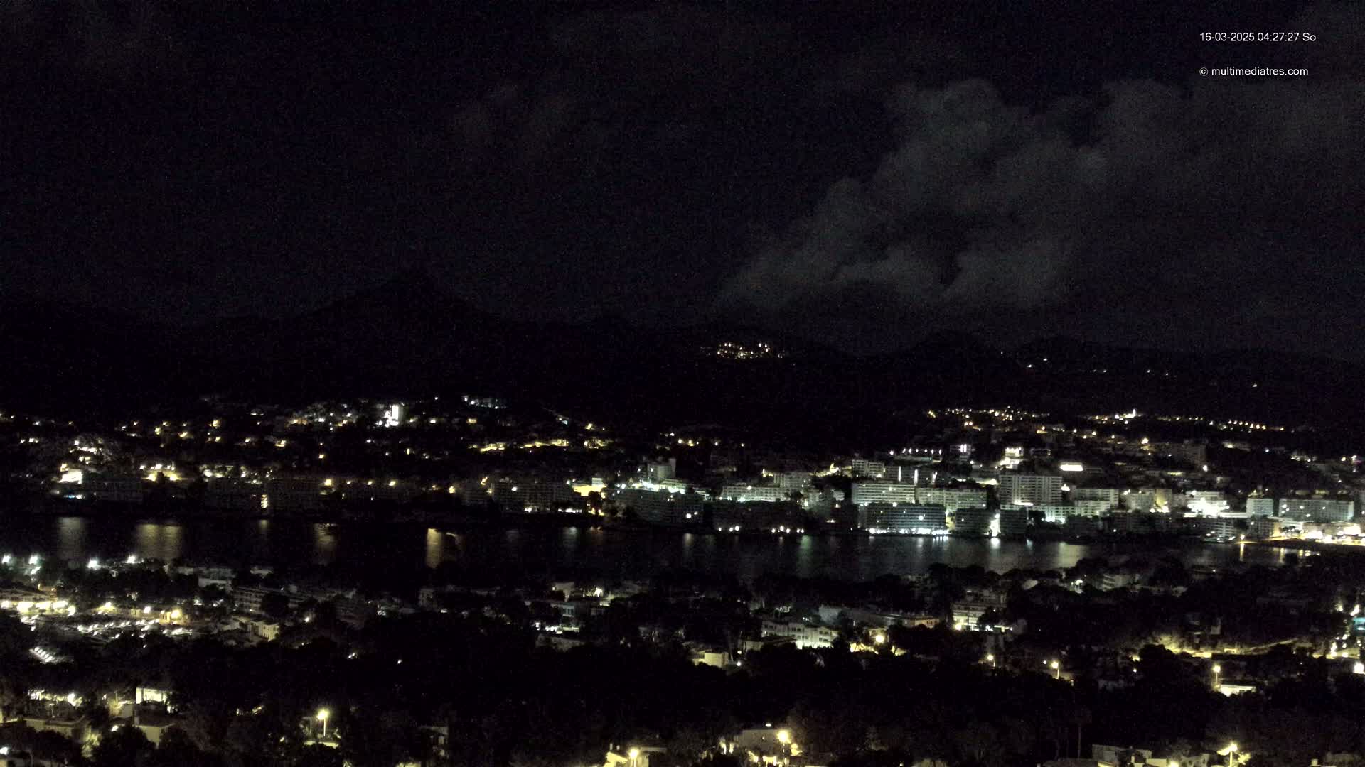 A nighttime, high-angle view of a coastal town illuminated by scattered lights, situated below dark hills under a partly cloudy sky.