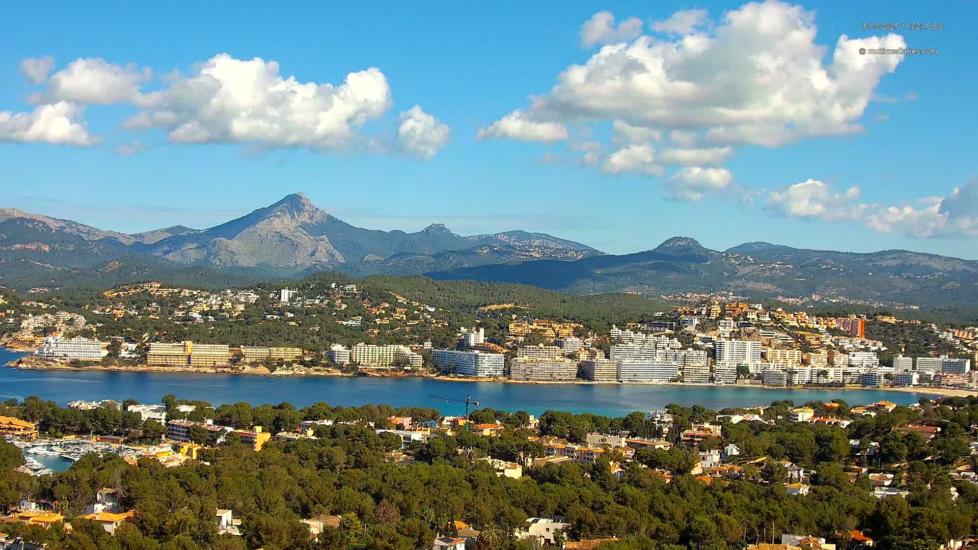 A coastal town nestled among green hills and mountains under a partly cloudy blue sky.