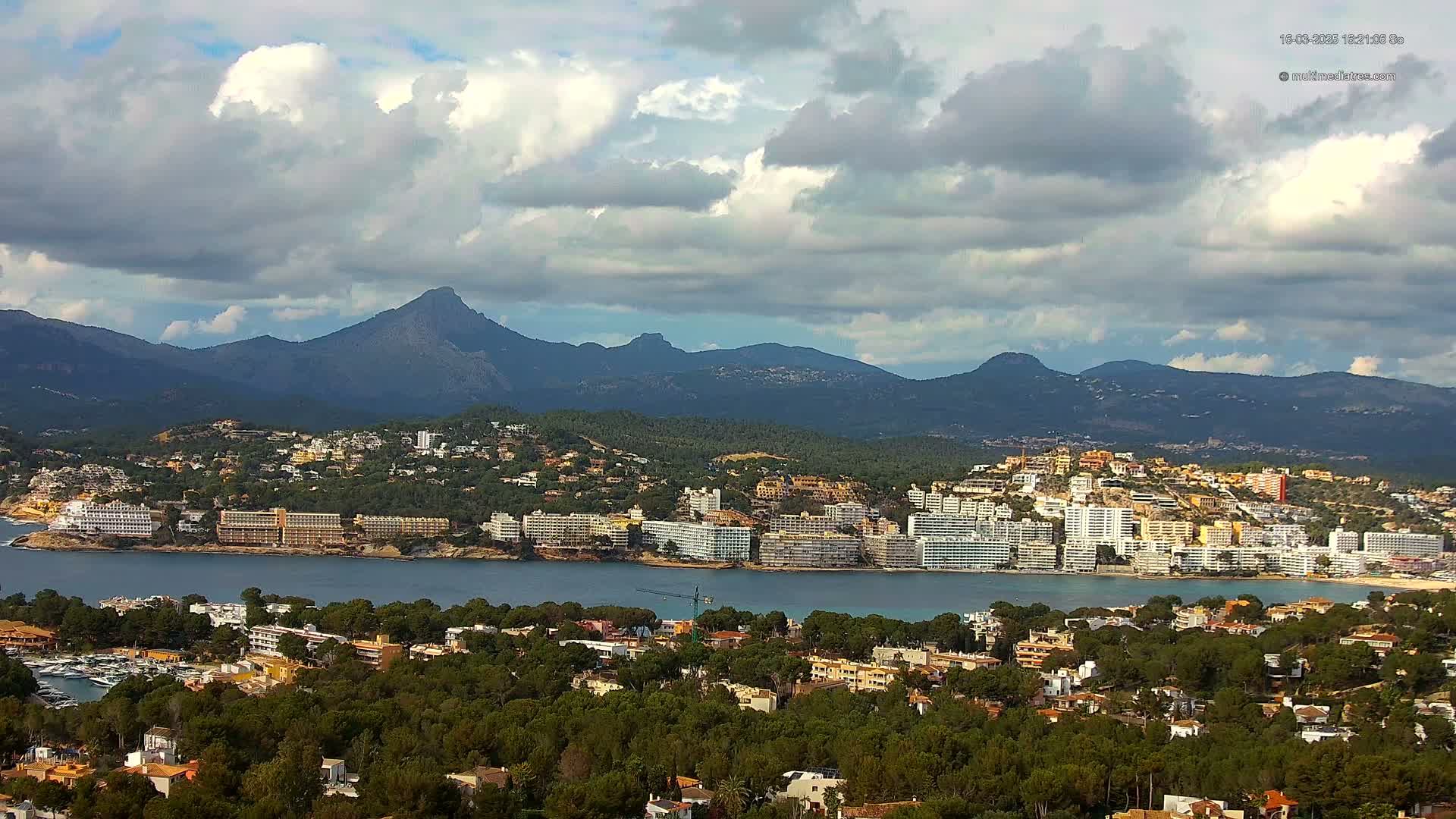 A coastal town nestled amongst green hills and mountains under a partly cloudy sky overlooks a calm bay.