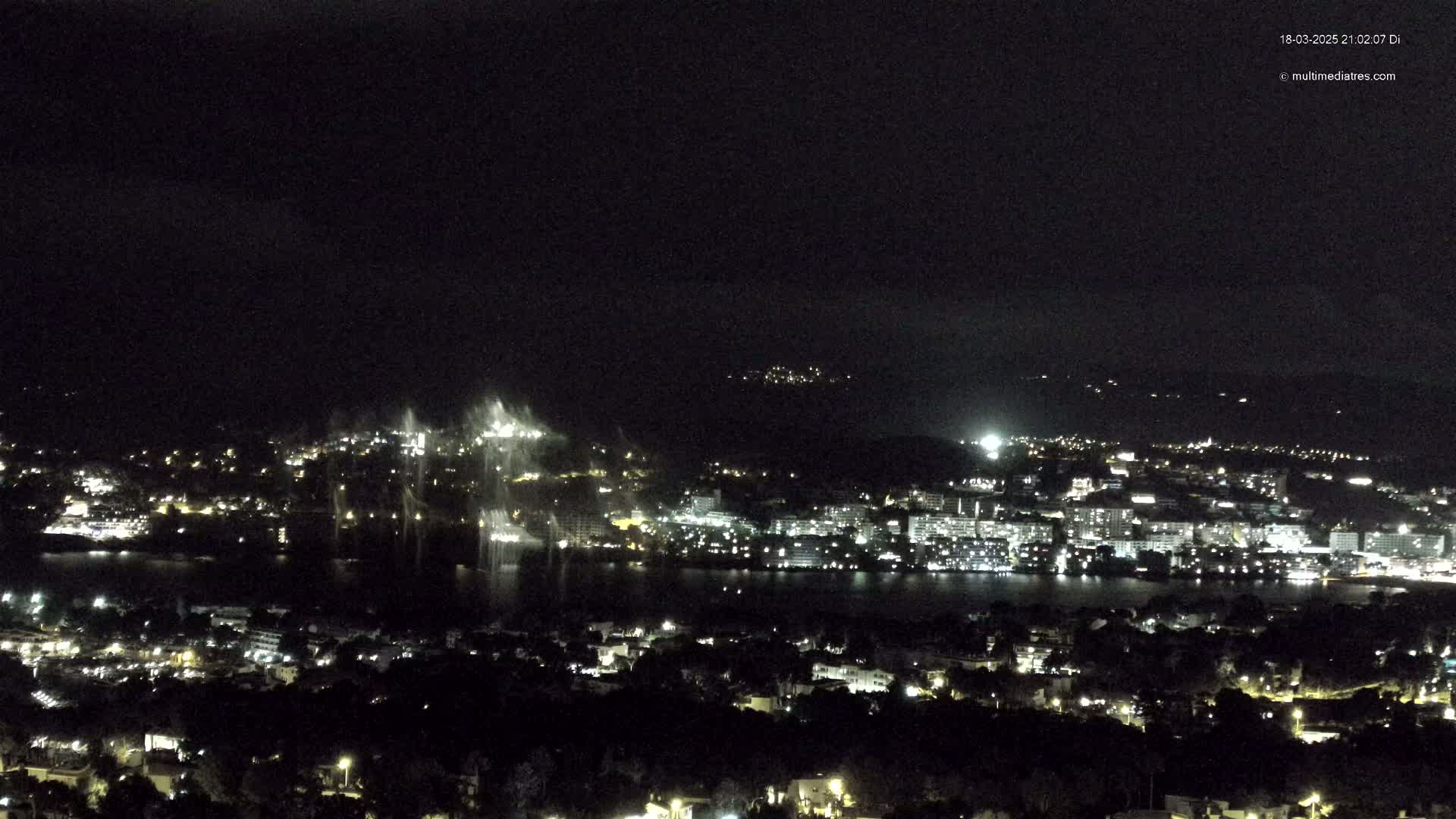 A nighttime view of a city and body of water, illuminated by lights, with a water fountain display visible in the distance under a dark sky.