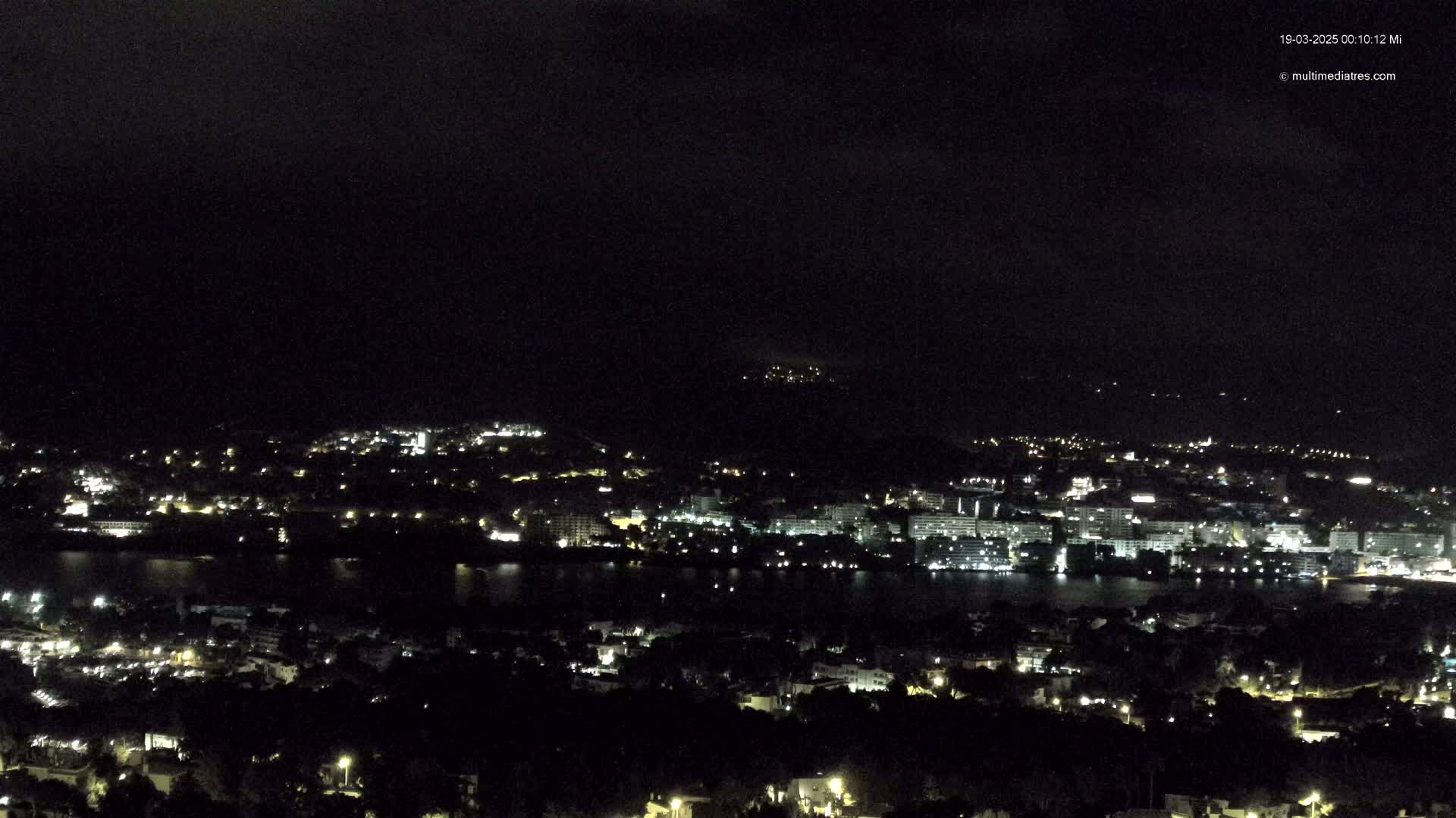 A nighttime, aerial view of a city and body of water, illuminated by scattered lights, under a dark, cloudy sky.