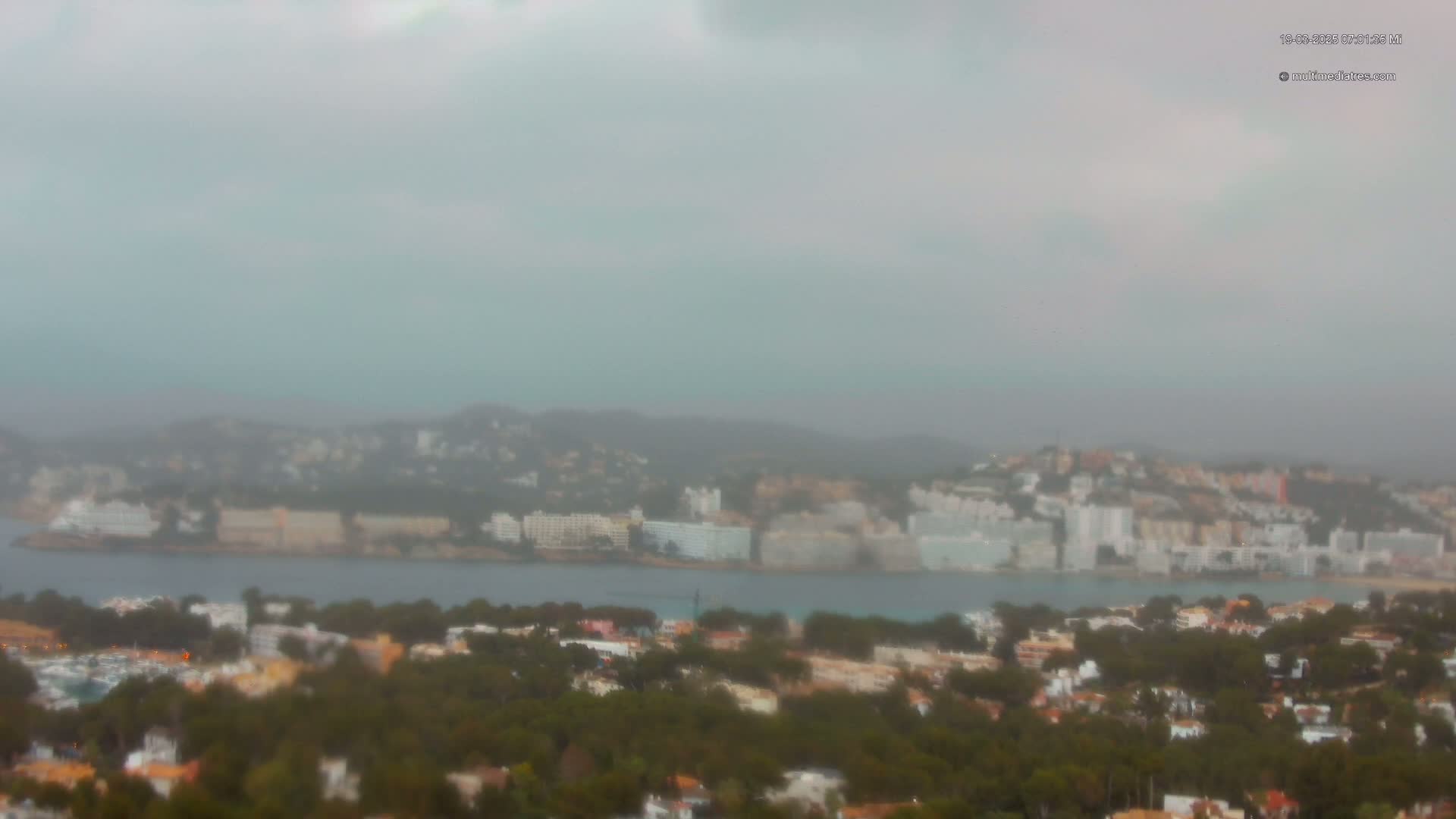 A hazy, overcast day reveals a coastal town nestled between a body of water and a hillside dotted with buildings.