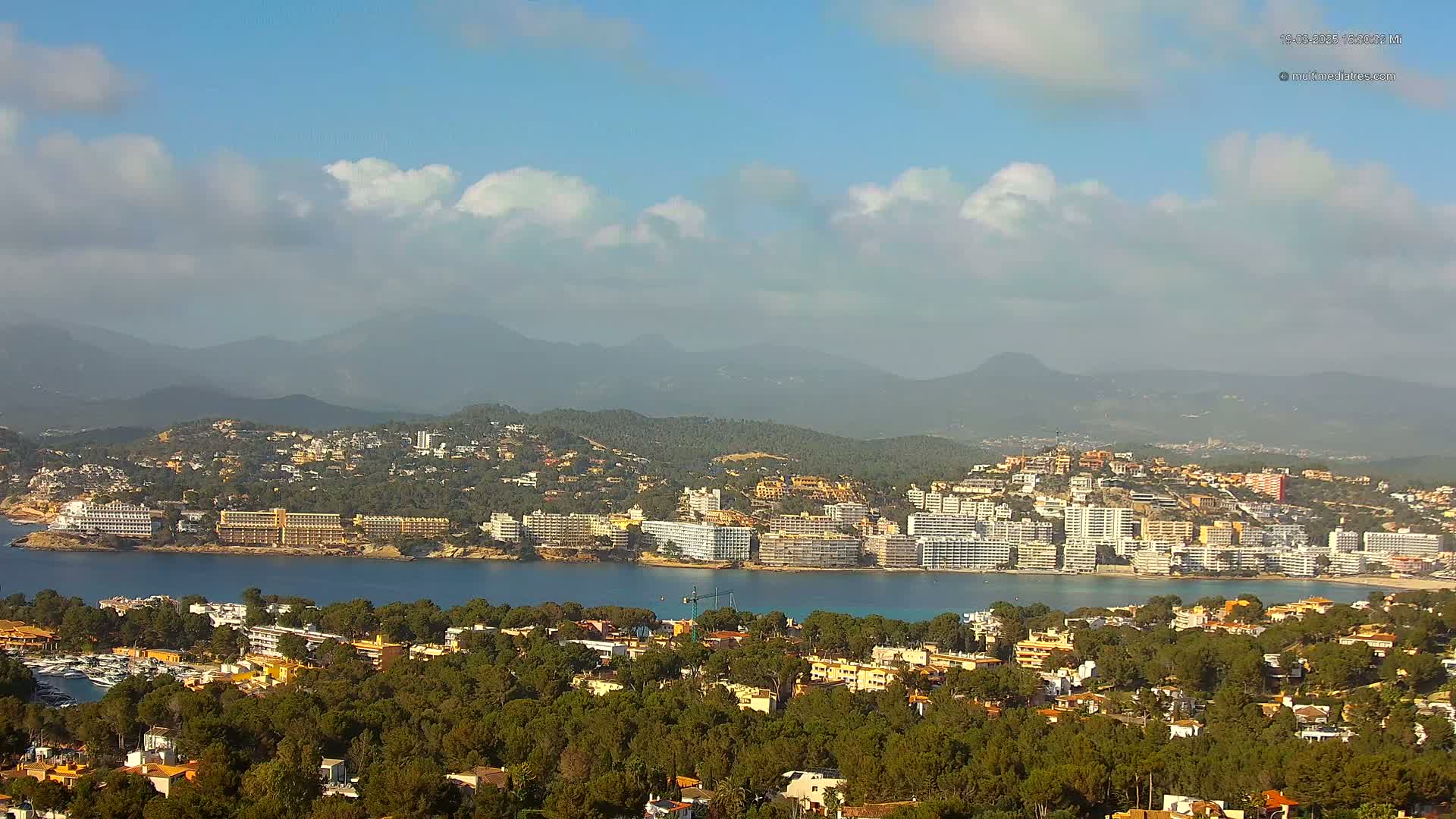 A coastal town with numerous hotels and buildings nestled amongst green trees, overlooking a calm bay, with hazy mountains in the background under a partly cloudy sky.