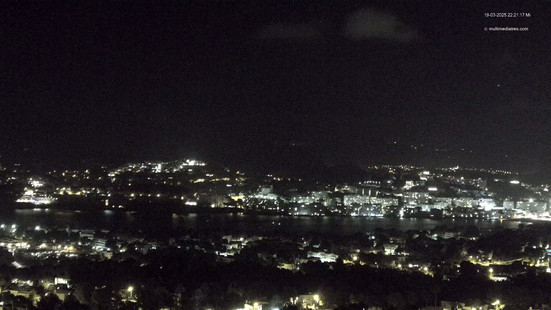 A nighttime, high-angle view shows a city with lights illuminating buildings along a dark body of water under a mostly clear, dark sky.