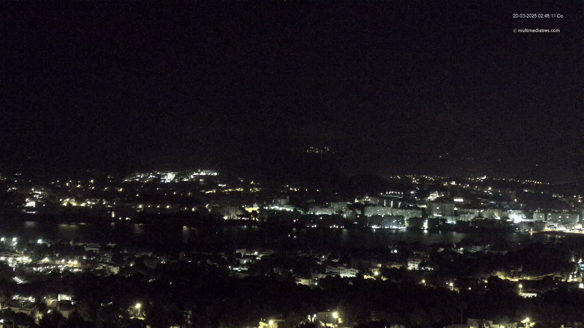 A nighttime, long-range view of a city and body of water illuminated by lights, under a clear, dark sky.