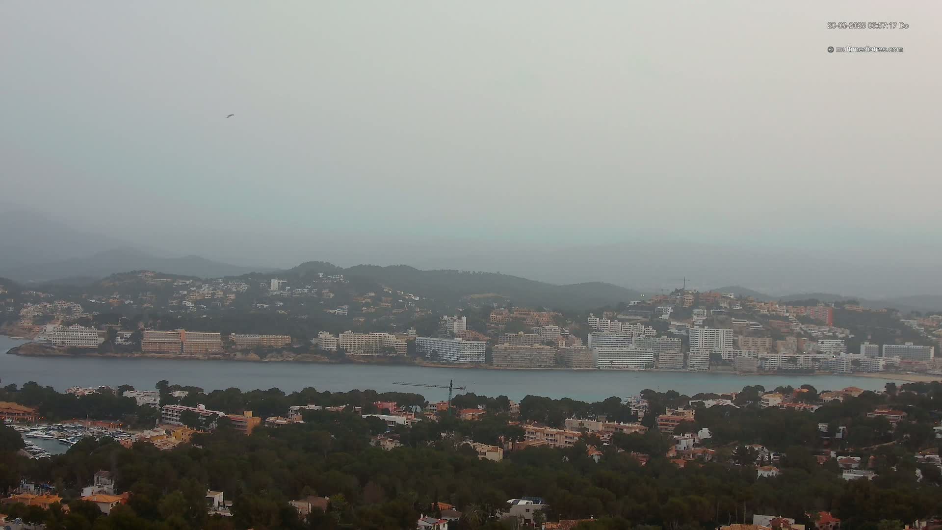 A hazy coastal town with many buildings and hotels sits on a hillside overlooking a calm body of water.