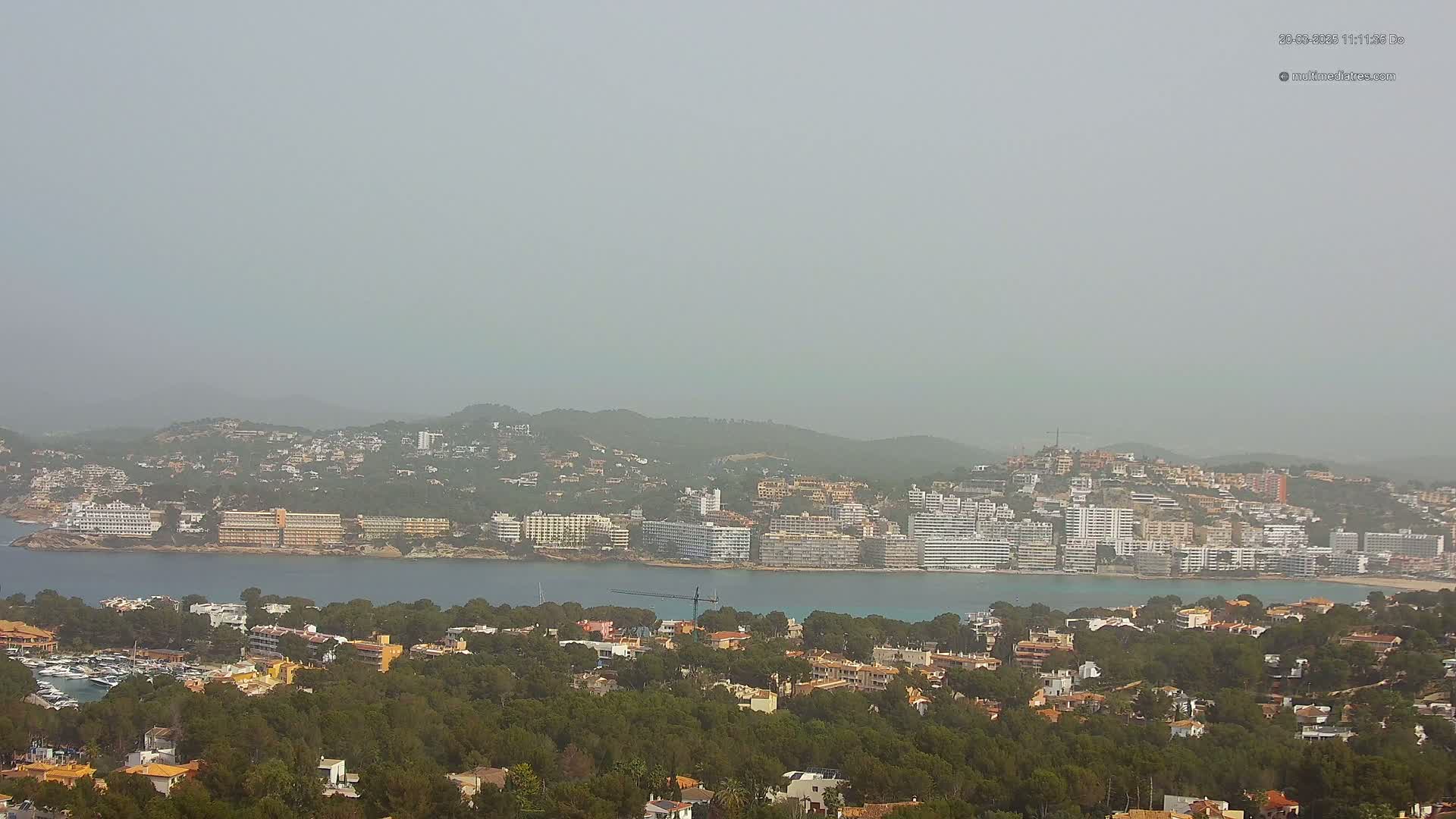 A hazy coastal town with numerous buildings situated on a hillside overlooking a calm bay, surrounded by green vegetation.