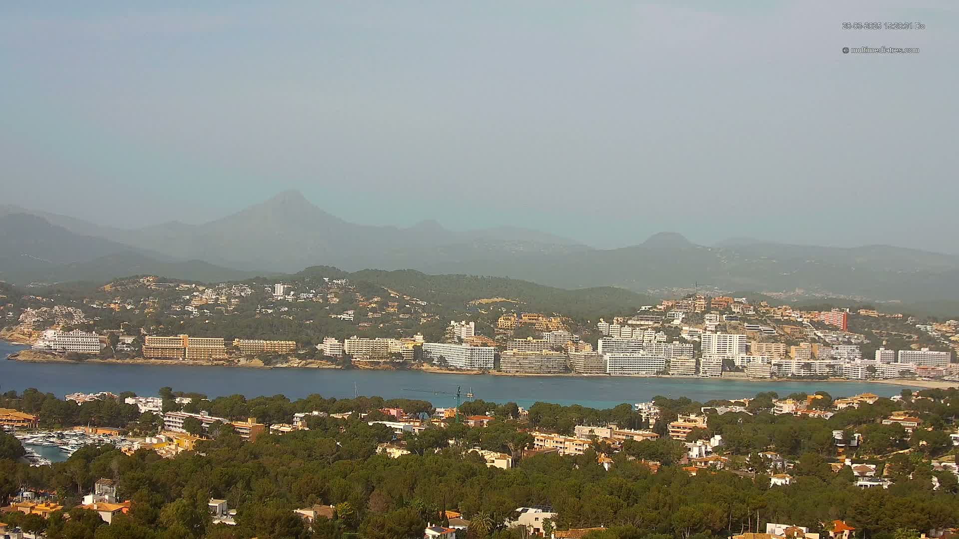 A hazy coastal town with numerous buildings and a marina sits nestled among green hills and mountains under a light-grey sky.