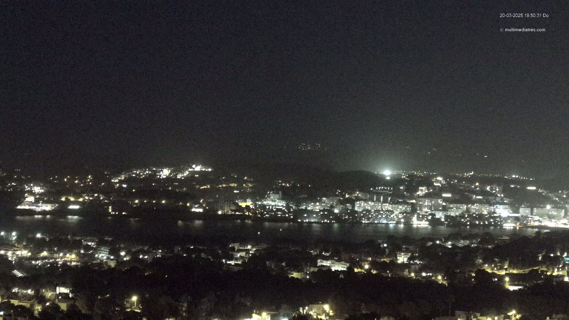 A nighttime view shows a city sprawling across a hillside, with its lights reflecting on a dark body of water, under a clear, dark sky.