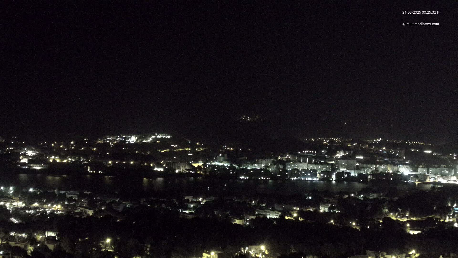 A nighttime, aerial view shows a city with lights scattered across hills and along a body of water under a clear, dark sky.