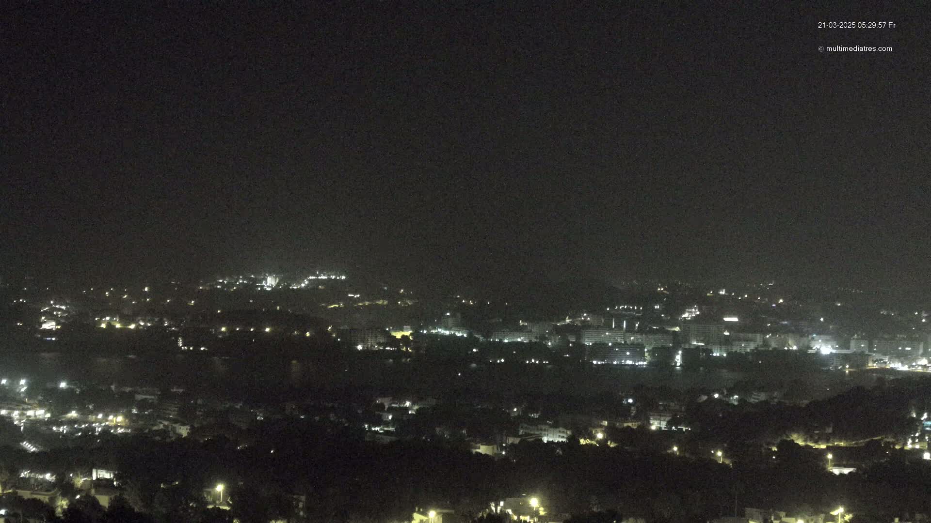 A nighttime long shot of a city and surrounding hills illuminated by scattered lights under a dark, clear sky.