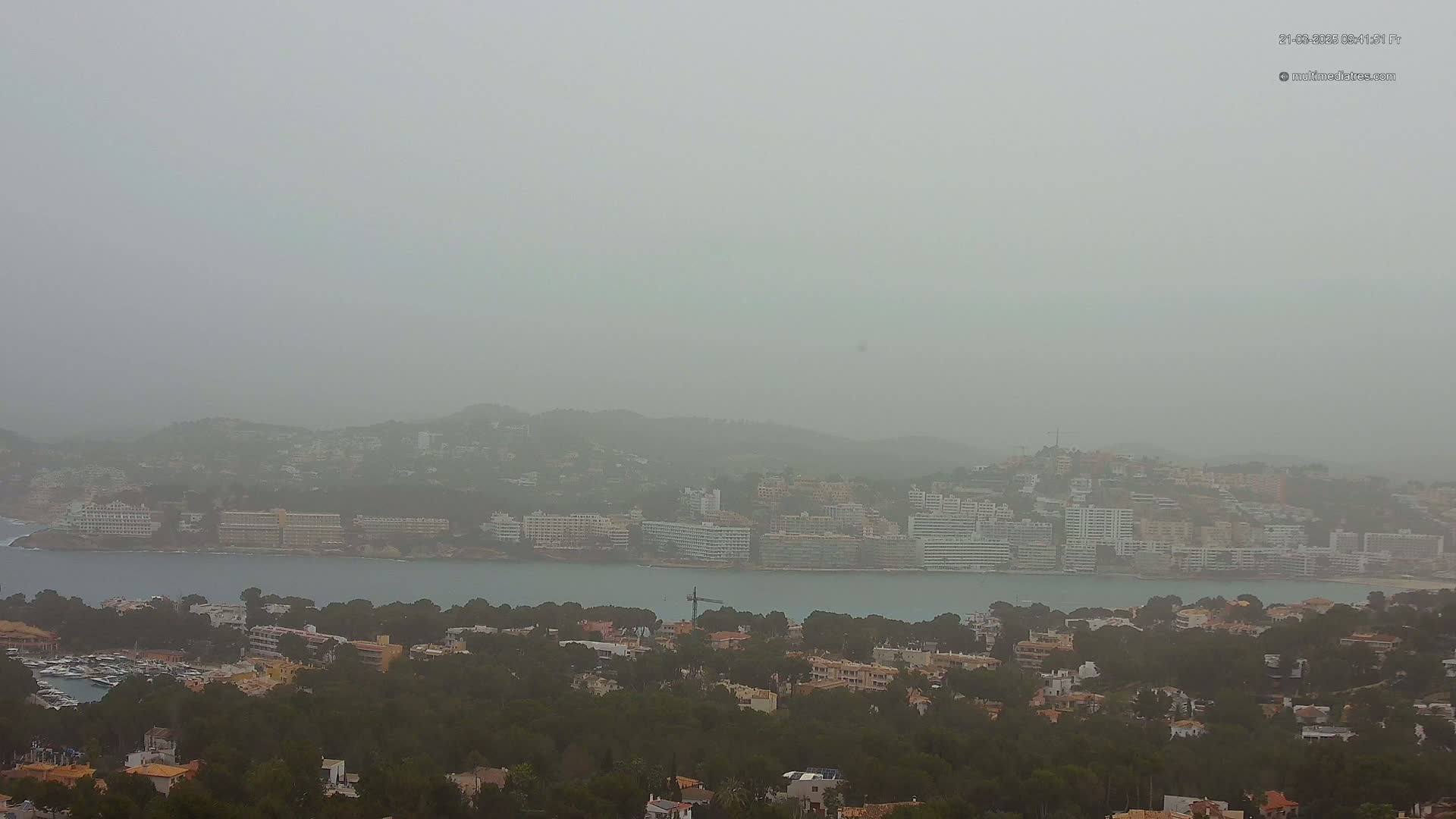 A hazy coastal town with numerous buildings is seen from an elevated vantage point in dusty or foggy conditions.
