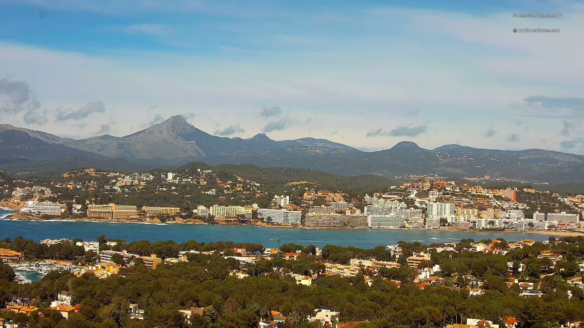A coastal town nestled between a bay and a mountain range under a partly cloudy sky.