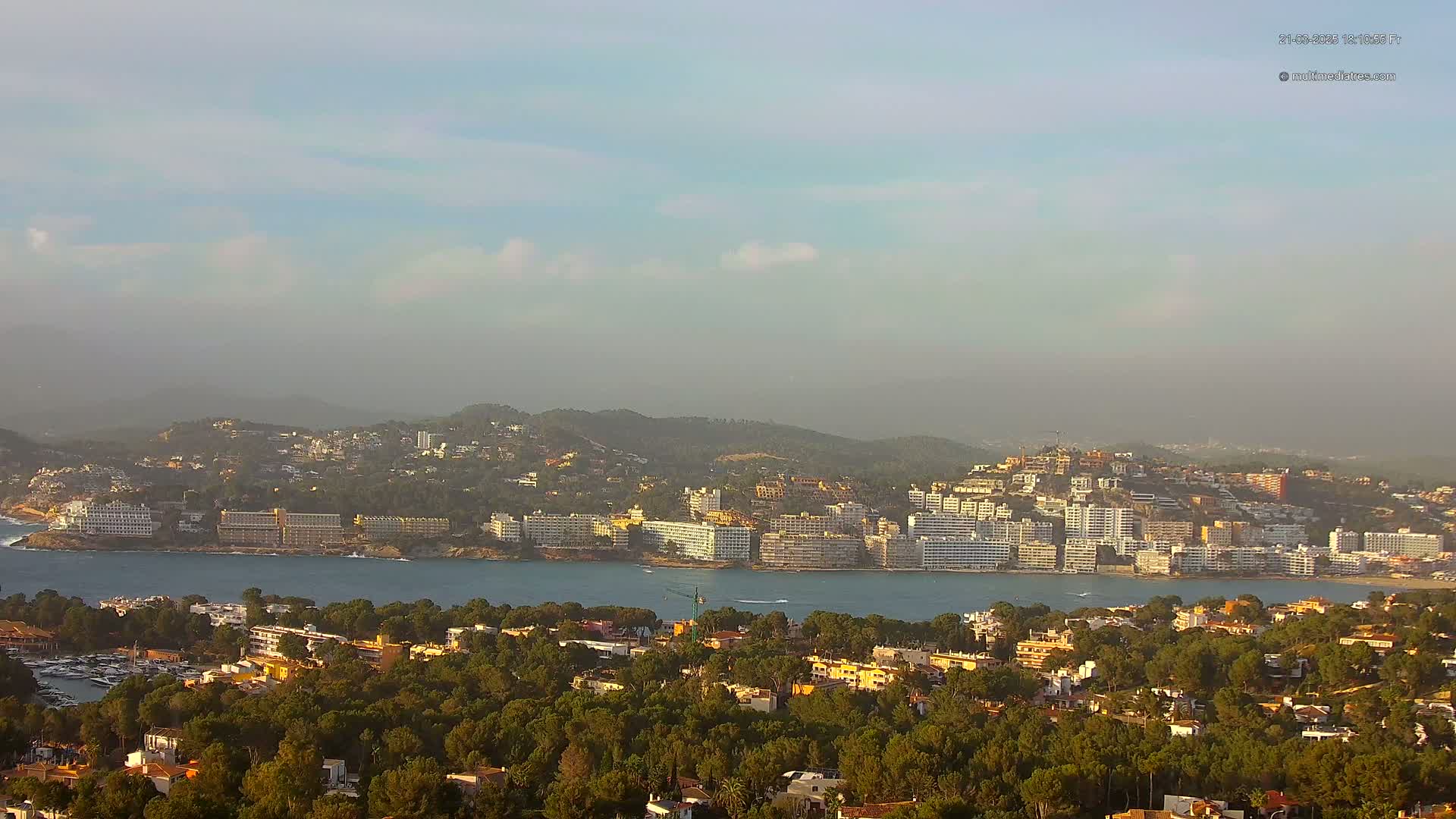 A hazy coastal town with many buildings, nestled between a calm bay and verdant hills.