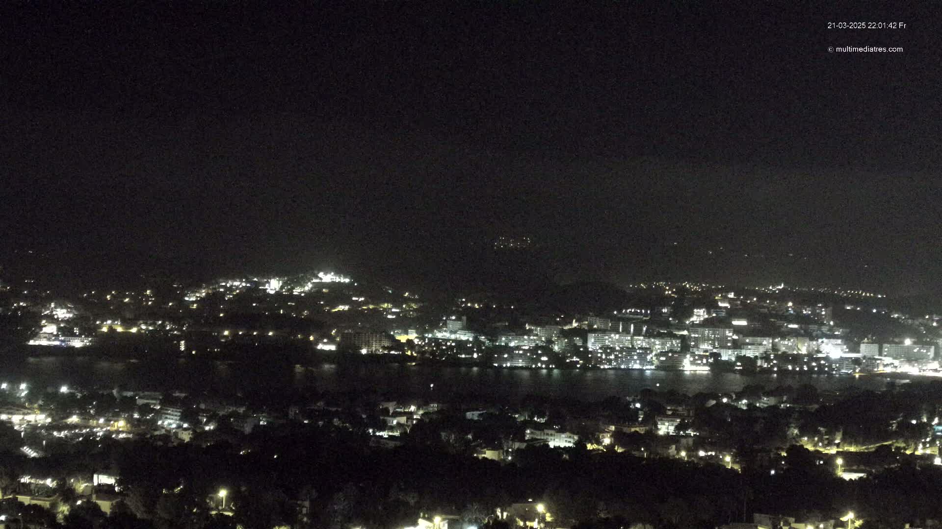 A nighttime view shows a city illuminated by lights along a dark body of water, under a dark, clear sky.
