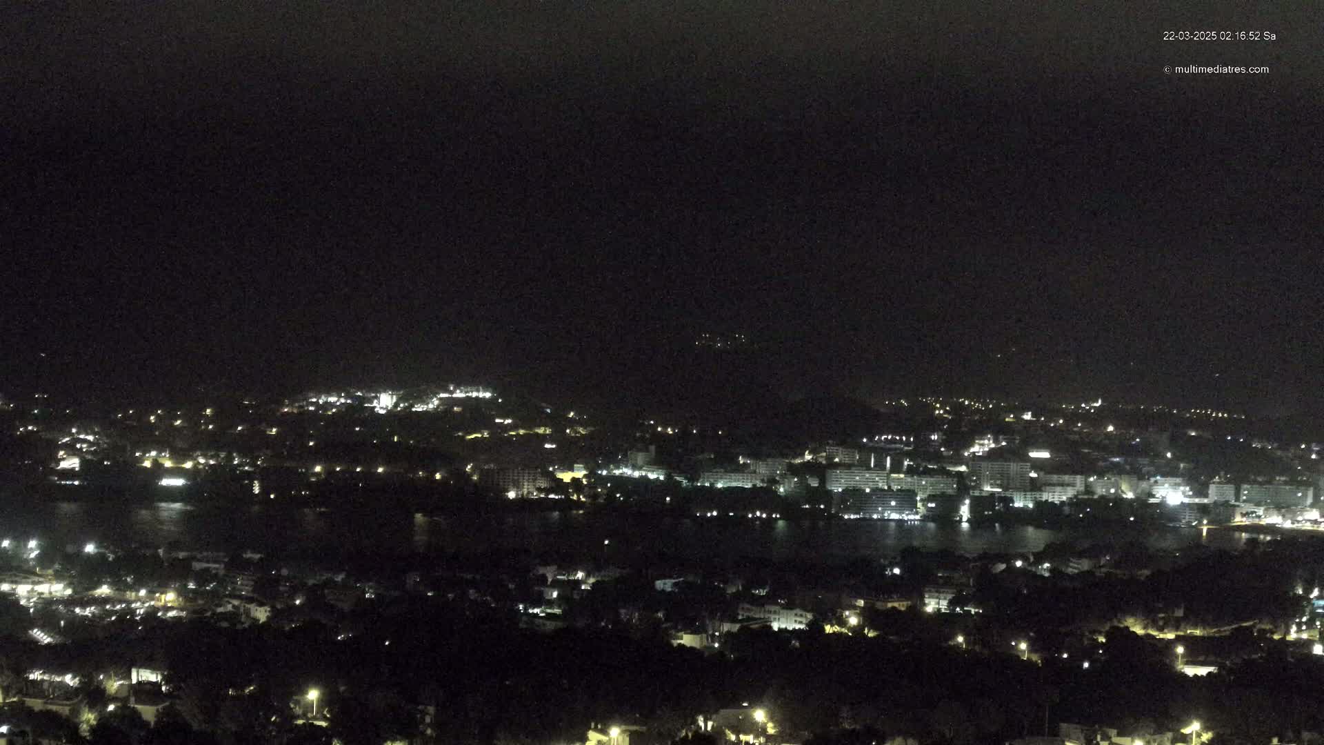 A nighttime, aerial view shows a city with many lights surrounding a dark body of water, under a clear, dark sky.